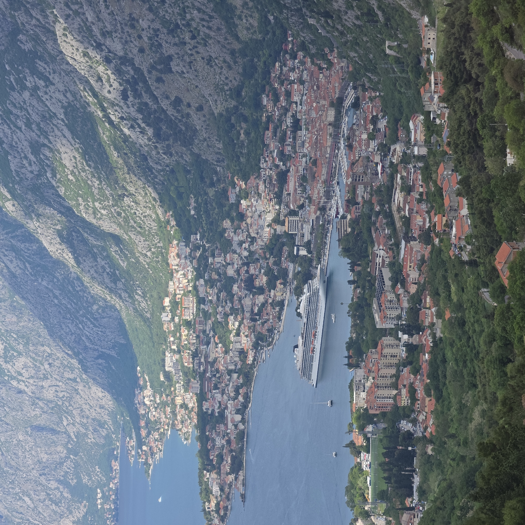 The image depicts an aerial view of Kotor, Montenegro, featuring the Bay of Kotor, a cruise ship docked in the bay, and surrounding mountains and buildings.
The scene includes:
1. The Bay of Kotor, a large body of water with a narrow inlet.
2. A large cruise ship docked near the town, indicating a tourist presence.
3. The old town of Kotor, characterized by dense housing with red-tiled roofs.
4. Mountains on either side of the bay, with visible texture and vegetation.
5. Buildings of various sizes, from single-family homes to larger apartment complexes.
6. Roads running along the shoreline.
7. Vegetation including trees and shrubs.
8. Some boats moving on the water.
9. A possible soccer field.
10. Houses and buildings further into the forested mountain slopes.