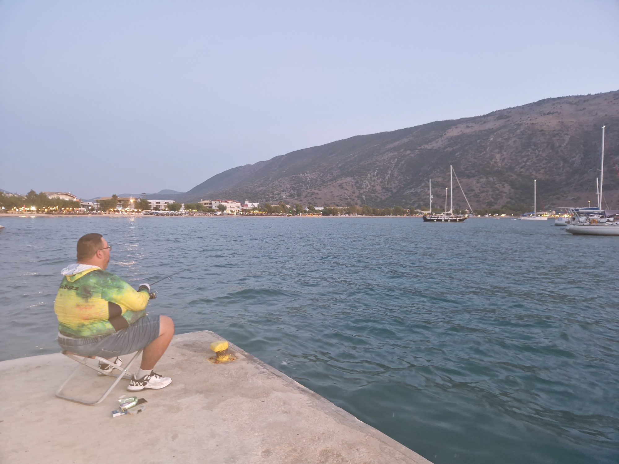 A man is fishing from a concrete pier in Plataria, Greece, with several sailboats in the water in the background. The man is sitting on a small folding chair, wearing a brightly colored hooded shirt and denim shorts. He is holding a fishing rod and appears to be casting his line into the water. The pier is made of concrete and has a yellow bollard on it. The water is a deep blue and appears to be choppy. In the background, there are several sailboats moored, and a coastal town with buildings and trees can be seen along the shoreline. A large, tree-covered hill rises behind the town. The sky is a pale blue and appears to be twilight.