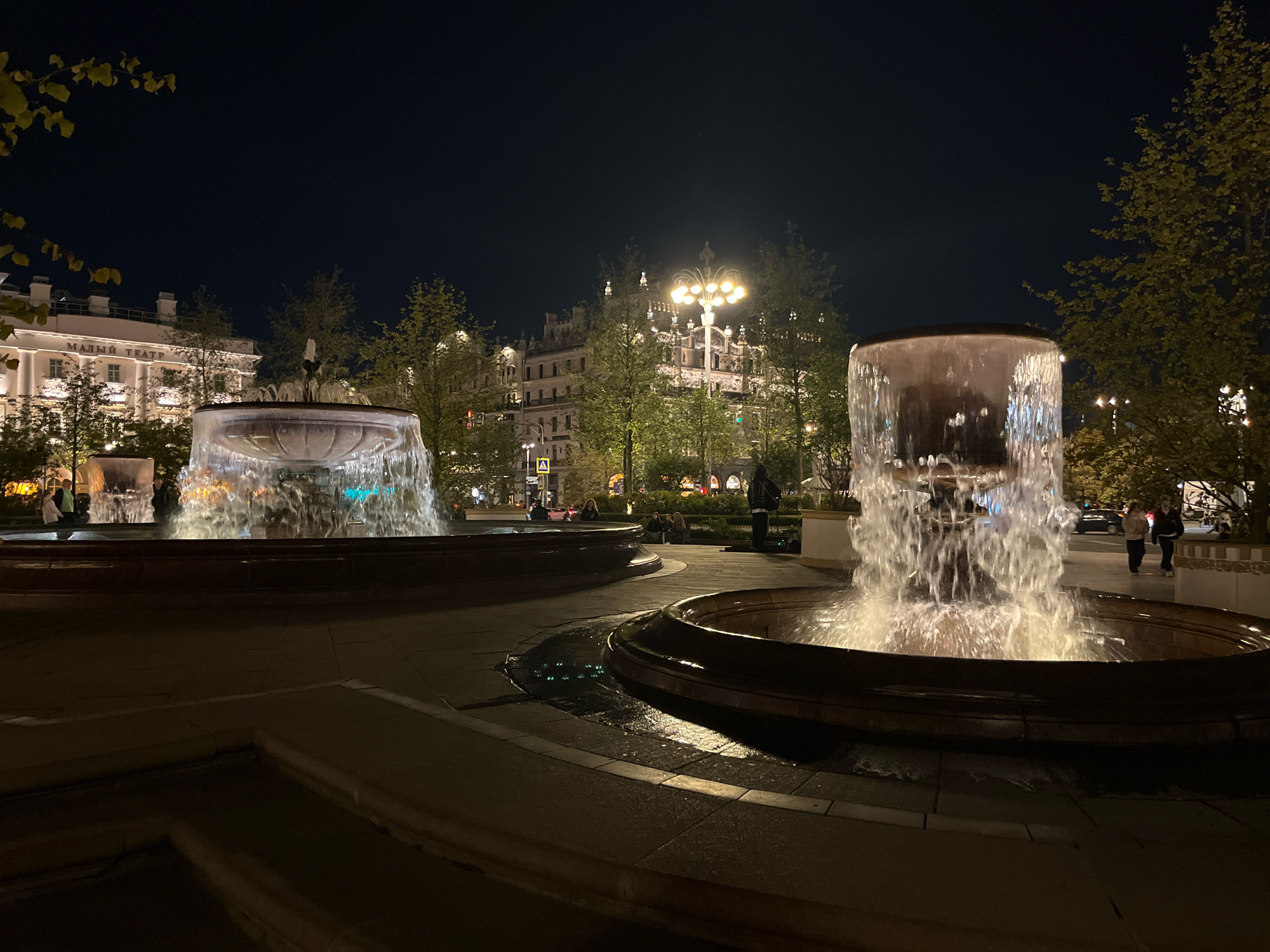 This is a nighttime photograph of fountains in a city square. In the foreground, two large, multi-tiered fountains are in operation, with water cascading down their sides and illuminated from within. The fountain on the right is closer to the viewer and its water flow is highly detailed. In the midground, trees line a walkway, and behind them are several ornate buildings with illuminated windows and architectural details. The building on the left has text that reads "МАЛЫЙ ТЕАТР" (Maly Theatre). A street lamp with multiple bulbs is visible above the trees. A few people are visible in the midground, some walking and some seated. Cars are present on a street behind the fountains and trees. The overall environment suggests a well-lit, public urban space at night, likely in Moscow, Russia, given the text on the building.