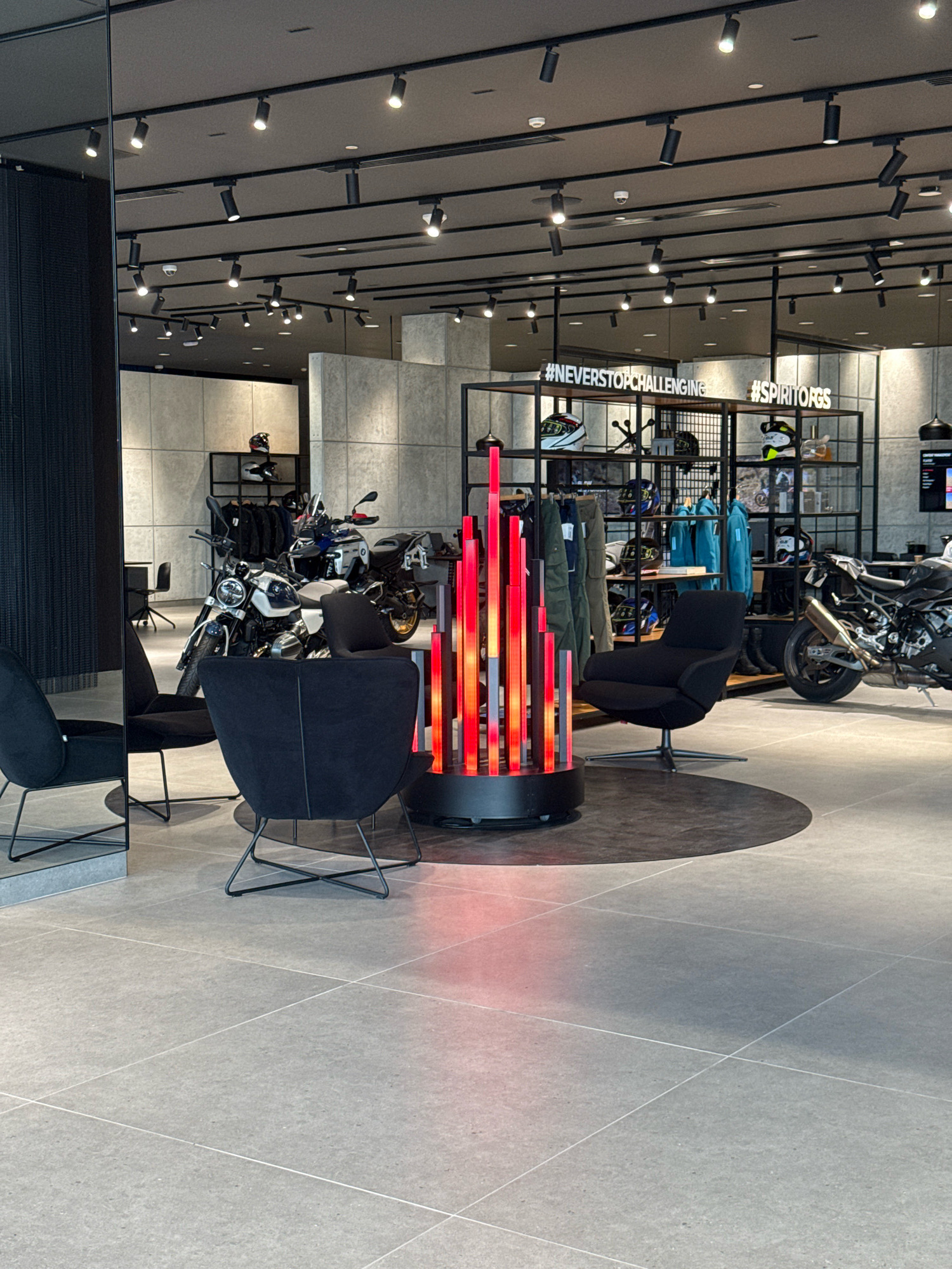 The image depicts the interior of a modern motorcycle dealership, likely a BMW Motorrad showroom, in Dubai, United Arab Emirates. The space is illuminated by a network of ceiling track lighting. In the foreground, three black upholstered armchairs with metal frames are arranged on a grey tiled floor with a circular dark grey rug in the center. A tall, vertical, red illuminated art installation is positioned between two of the chairs.
In the mid-ground, several motorcycles are displayed. A white and silver BMW R 1250 GS motorcycle is prominently featured, with another black motorcycle partially visible to its right. Behind the motorcycles, a black metal shelving unit displays various items, including helmets, apparel, and accessories. Two prominent signs on the shelving unit read "#NEVERSTOPCHALLENGING" and "#SPIRITOFGS".
The background reveals a concrete-textured wall and additional display areas. A mirrored pillar is visible on the left side of the image. The overall environment suggests a clean, upscale retail space designed to showcase motorcycles and related merchandise.