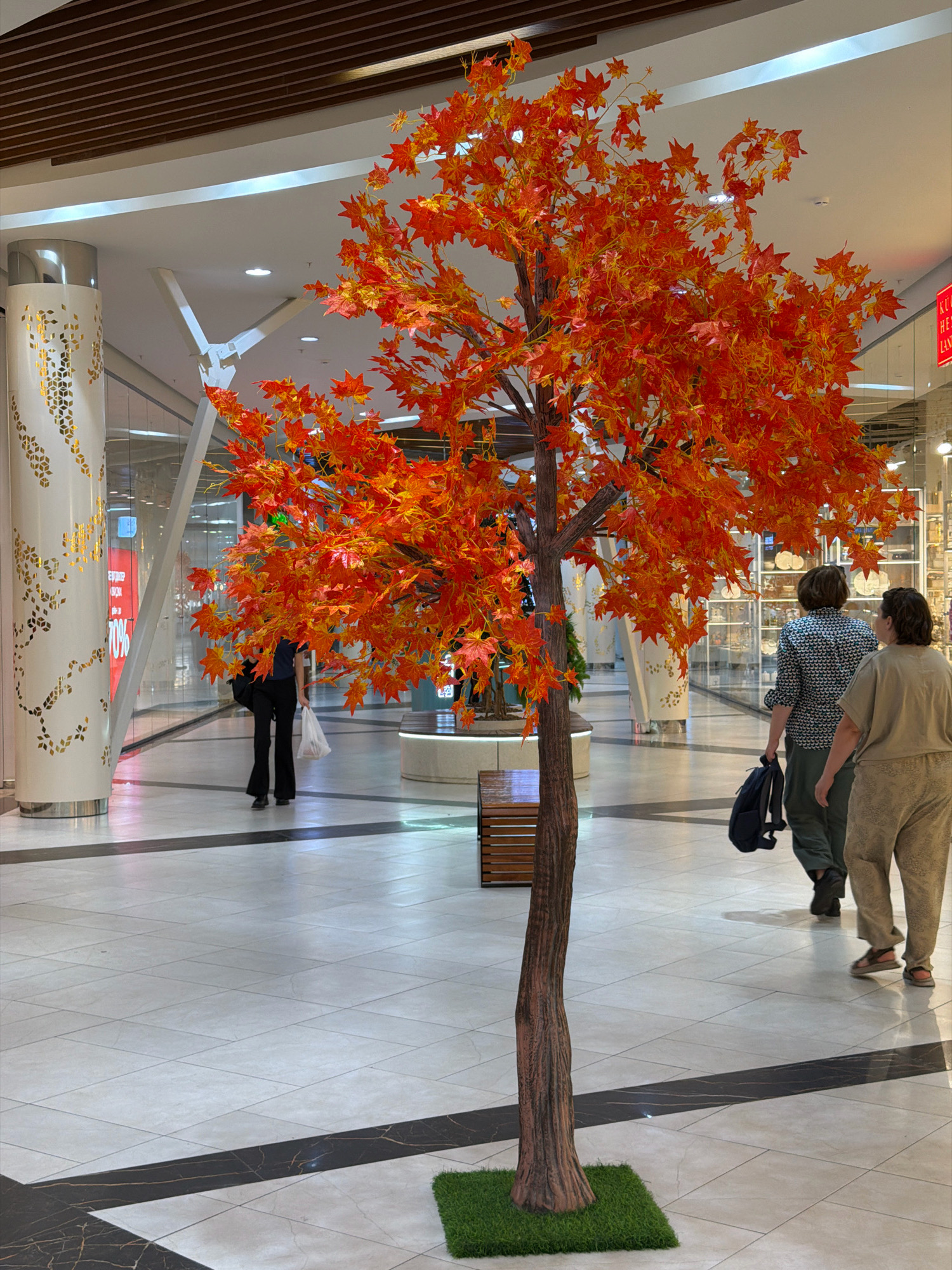 An artificial maple tree with bright orange and yellow leaves stands prominently in the center of a shopping mall. The tree has a textured brown trunk and is rooted in a square of artificial green grass. In the background, two women walk away from the camera. The woman on the left wears a patterned shirt and dark pants, carrying a dark backpack. The woman on the right is dressed in light-colored, loose-fitting clothing. To the left of the tree, a white cylindrical column with decorative gold geometric patterns is visible. The floor is tiled in a light beige color with dark accent lines. The ceiling of the mall features wooden slats and modern lighting. In the far background, shop displays and other shoppers can be seen. The scene appears to be a public indoor space, likely a shopping mall in Pavlodar, Kazakhstan, given the context.