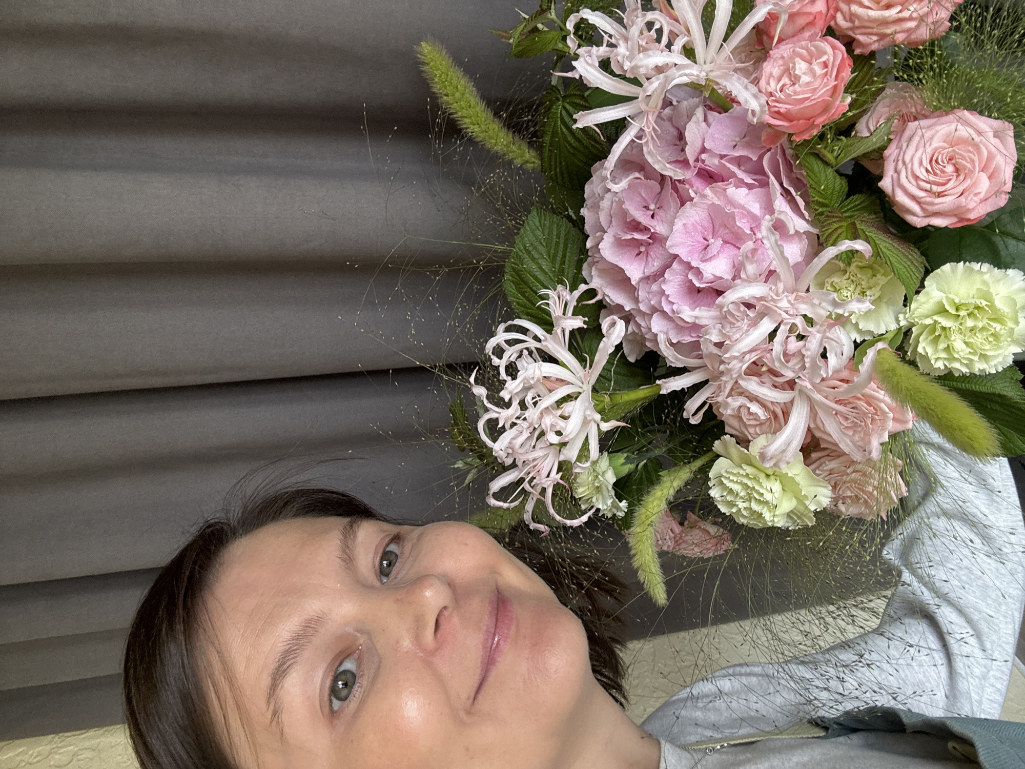 An overhead, medium shot captures a woman holding a bouquet of flowers. The woman, positioned on the left side of the frame, is looking at the camera and smiling. She has short brown hair, light skin, and is wearing a gray sweatshirt. The bouquet, filling the right side of the frame, consists of pink hydrangeas, pink roses, light green carnations, and delicate pink spider lilies, interspersed with thin, wispy grass. The background is a plain gray curtain. The image appears to have been taken indoors. There is no contextual information suggesting a location in Izhevsk, Russia.