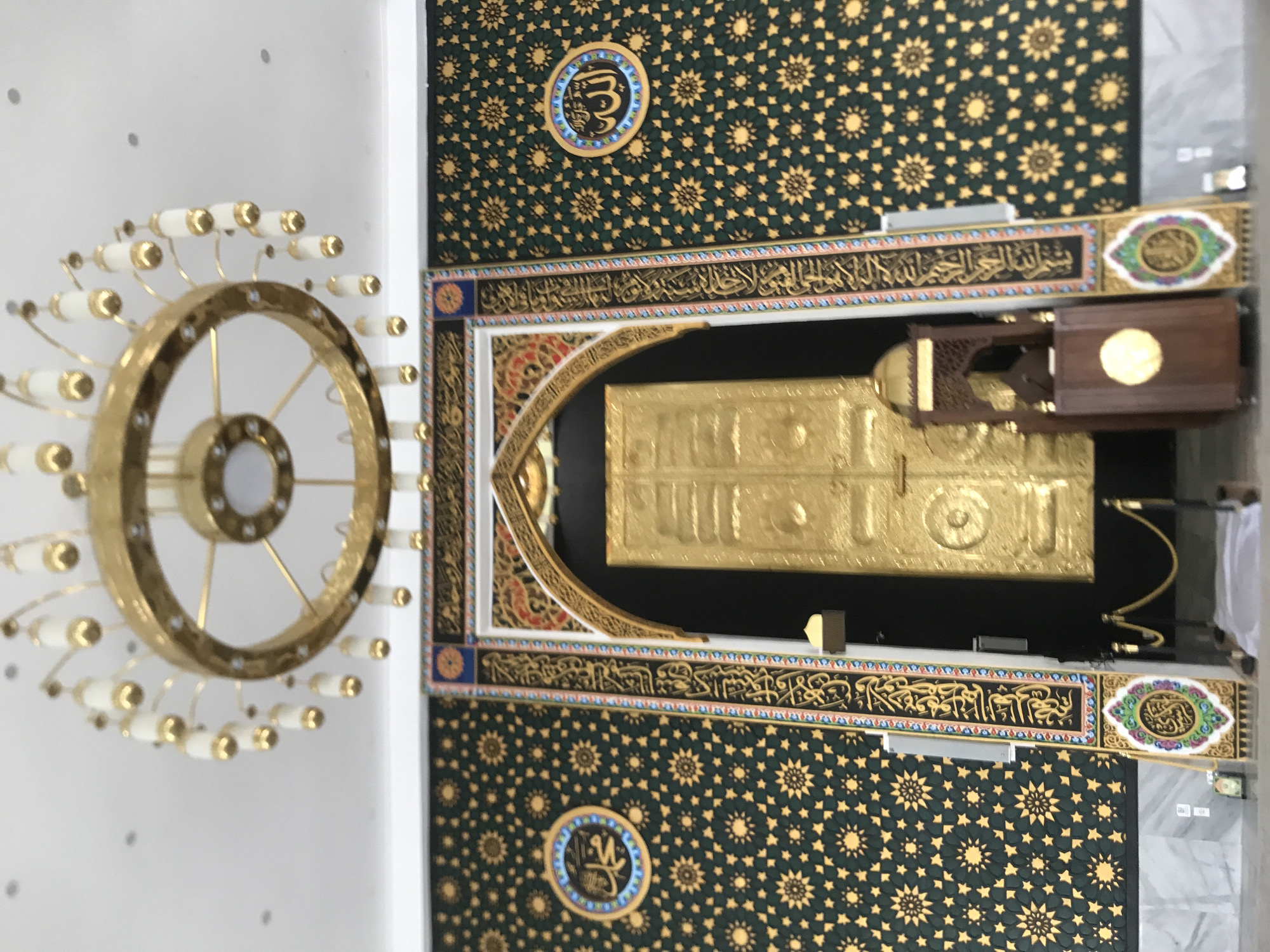 The image shows an interior view featuring a model or representation of the Kaaba, a central religious site in Islam. The Kaaba is a gold, cube-shaped structure with a black cloth covering, elaborately decorated with gold and Arabic calligraphy. It's positioned against a background of green patterned wallpaper with gold star-like motifs.
Above the Kaaba, there's a band of Arabic calligraphy. To the left of the Kaaba, a large circular chandelier with multiple lights hangs from the white ceiling.
The overall environment appears to be a building or exhibition hall. The location provided is Meunasahaluedrienlhoksukon Barat, Indonesia.