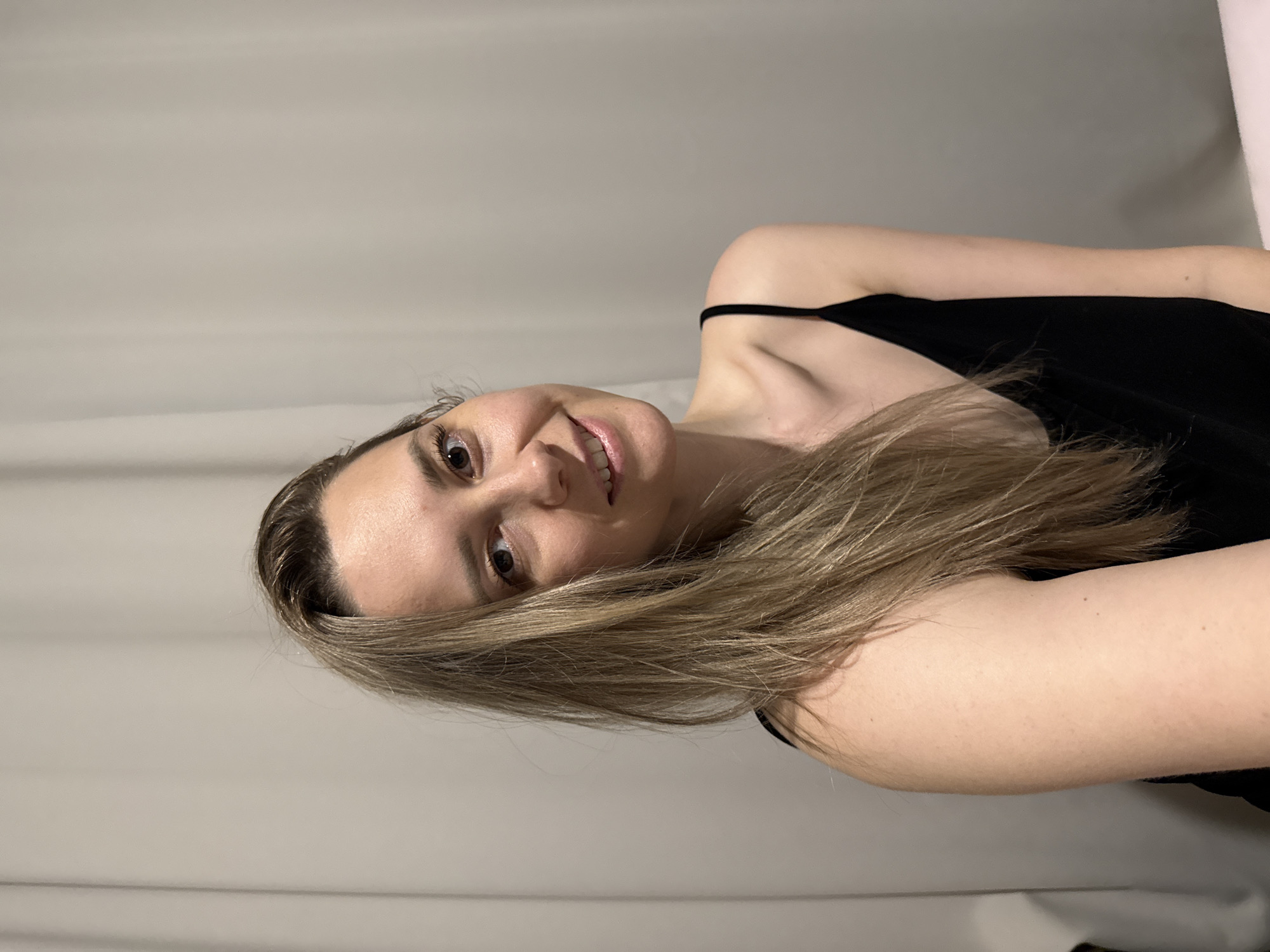 A woman with light brown hair is smiling at the camera. She is wearing a black tank top. The background is a plain, light-colored wall. The image does not provide any information to determine the location in Chaikovsky, Russia.