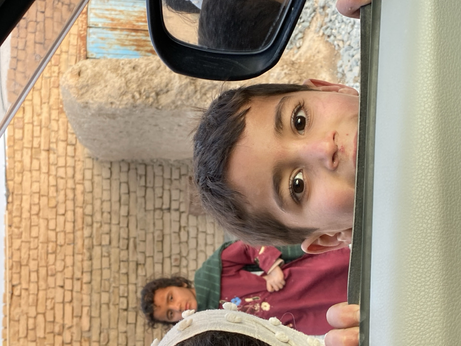 The image depicts an outdoor scene in Kandahar, Afghanistan, captured from the interior perspective of a vehicle looking outward. In the immediate foreground, a young child, appearing male, with dark hair and brown eyes, peers over the light-colored, textured car door frame, with his hands gripping the top edge. A small mark is present on his philtrum.
To the left and slightly behind, a partial view of a person wearing a light-colored, textured head covering is visible. Further back, a female child with dark, curly hair stands, wearing a maroon long-sleeved dress with embroidery details and a dark green shawl draped over her shoulders; her left hand is placed on her hip.
The background consists of a light-brown brick wall. To the right of the wall, a tall, irregular, light-brown adobe structure is visible. Further right, a partially rusted blue metal gate or door is present. The ground in the distance, and as a blurry reflection in the black car side mirror on the far right, appears to be gravel and dirt. The sky is indistinct and light.