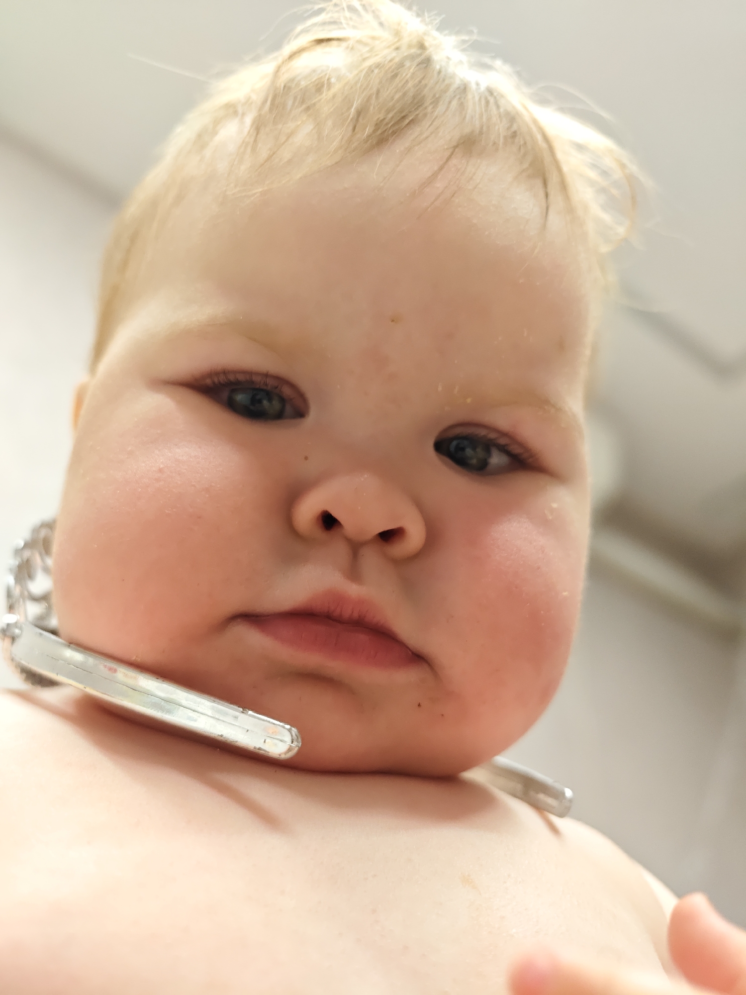 A close-up, low-angle portrait captures a baby with a serious or contemplative expression, looking slightly downward. The baby has fair skin, chubby cheeks, and light blonde or light brown hair that appears fine and slightly disheveled on the forehead. Its dark eyes are open and focused.
A prominent metallic object rests around the baby's neck and upper chest. This object appears to be silver-colored and has two distinct parts: a chain-link section visible on the left side of the baby's neck, and a more solid, straight, shiny bar or plate-like section extending across the right side of the chest, resting on the skin. This object could be a large piece of jewelry, a toy chain, or an accessory.
The background is uniformly light-colored and out of focus, suggesting an indoor setting with diffused or artificial lighting. There are no specific distinguishing features in the background to indicate a precise location, other than it being an interior space. Minor details on the baby's skin include a few small marks or blemishes on the cheeks and around the nose, and some light, flaky texture on the forehead. The image is a tight crop on the baby's face and upper body, emphasizing its features and the metallic item. No visible text is present.