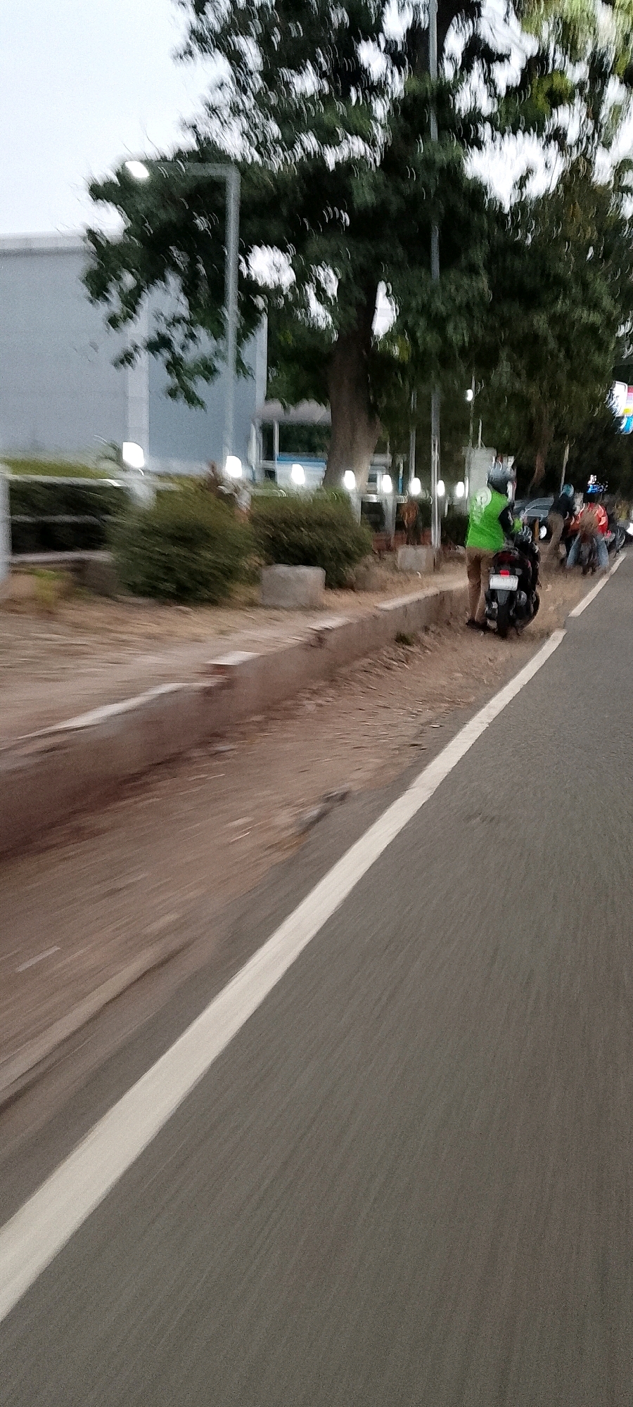 The image captures a street scene in Depok, Indonesia, with a focus on a person on a motorcycle. The perspective is from a moving vehicle, creating motion blur on the road and surroundings. In the foreground, a gray asphalt road with a white dividing line is visible, exhibiting motion blur. To the left of the road, a dirt embankment with some dry vegetation runs alongside. Further left, there is a low concrete barrier, and beyond that, a building with a gray facade. In the background, several trees with dense green foliage are present, interspersed with streetlights that appear as bright white orbs due to the motion blur.
The main subject is a person wearing a bright green jacket and a helmet, standing next to a dark-colored motorcycle. The person appears to be either preparing to mount or dismount the motorcycle. The motorcycle has a white license plate. Behind this individual, other motorcycles and people are visible in the background, also with significant motion blur, suggesting they are also on the road or sidewalk. The overall lighting suggests it is either early morning or late afternoon. The specific location within Depok is not identifiable from the image details.