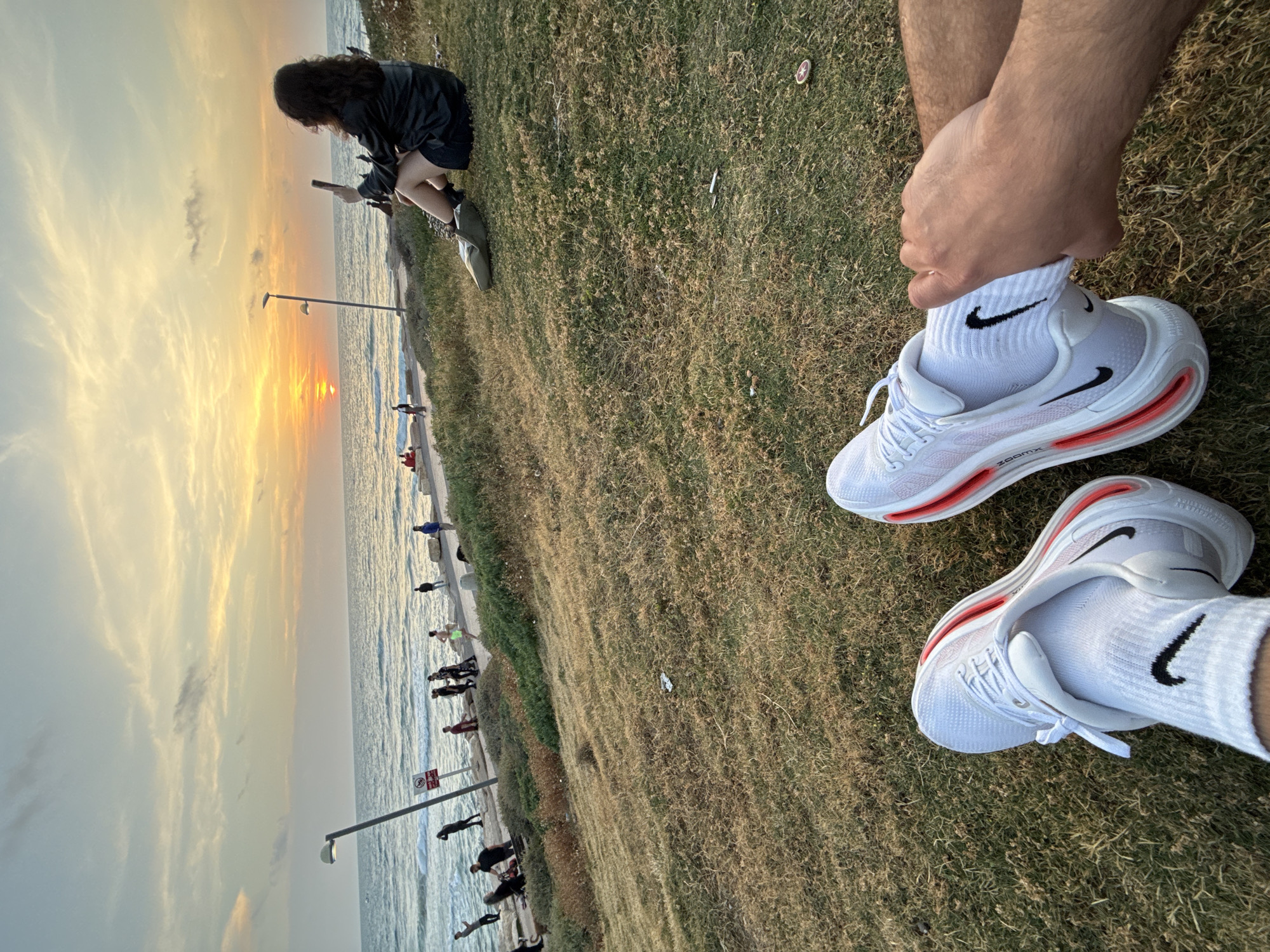 The image depicts a scene in Tel Aviv, Israel, during sunset. A grassy area overlooks a beach and the Mediterranean Sea, where people stroll along the coastline. A person wearing a black jacket and shorts is sitting on the grass looking at their phone. In the foreground, a person wearing white socks and Nike running shoes is visible. The image is taken from a high angle, focusing on the shoes and the sunset over the sea.