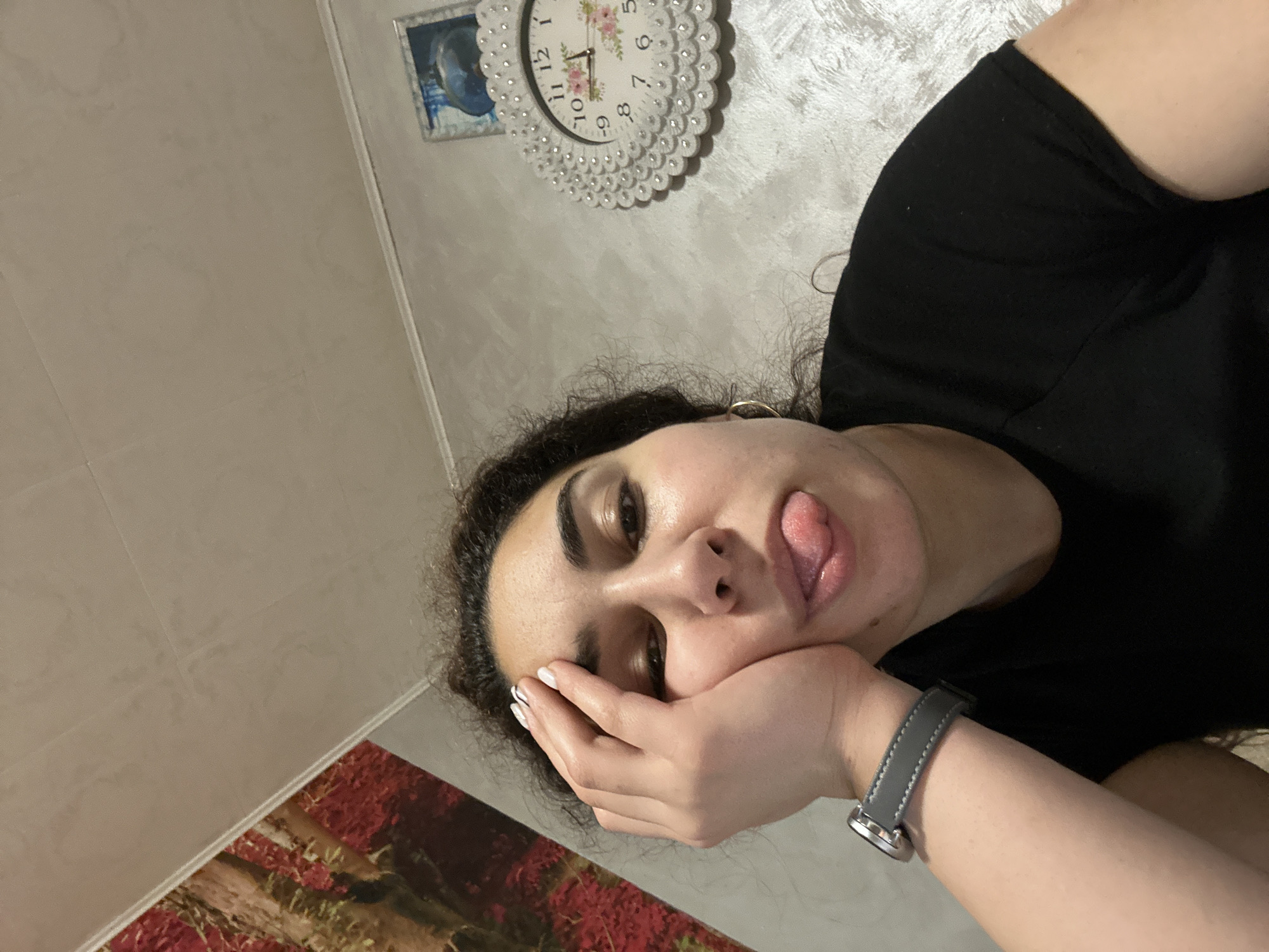 A young adult woman is lying down, sticking her tongue out with her face resting on her hand. She is wearing a black t-shirt and a watch with a gray band. The background includes a beige wall, a clock adorned with faux pearls, and a small framed image. A section of a patterned rug is visible at the bottom left. There is no indication of the location being Burgas, Bulgaria.