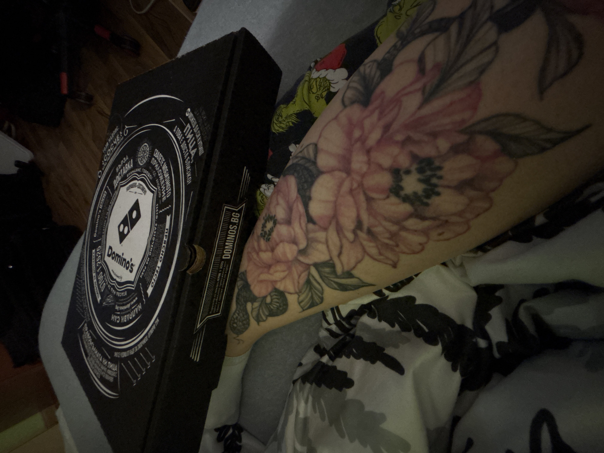 The image shows a Domino's pizza box alongside a person's arm with a floral tattoo. The box is predominantly black with a white circular logo and text in Bulgarian. The text on the box includes "Domino's," "Sofia," and a phone number. The person's arm is visible from the elbow down, covered in a floral tattoo. The background includes a patterned fabric that is likely a blanket or pajamas. The pizza box is the DOMINOS.BG franchise.