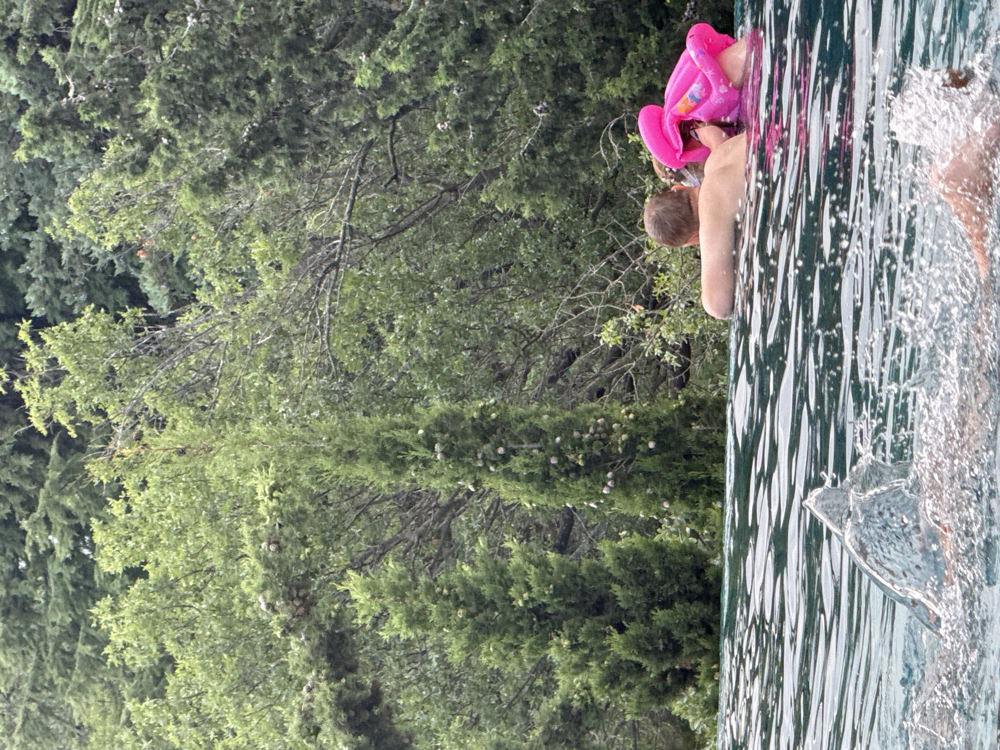 In the city of Yalta, Ukraine, two individuals are immersed in a swimming pool, their upper bodies visible above the water. The person closer to the foreground, from the back, has light brown hair and is wearing goggles. Beside this individual, a second person is partially obscured by a bright pink inflatable. The background is a dense wall of lush green trees, with the pool appearing to be an infinity pool, given the sharp drop-off visible at its edge. Water is splashing in the foreground, obscuring the lower portion of the scene.