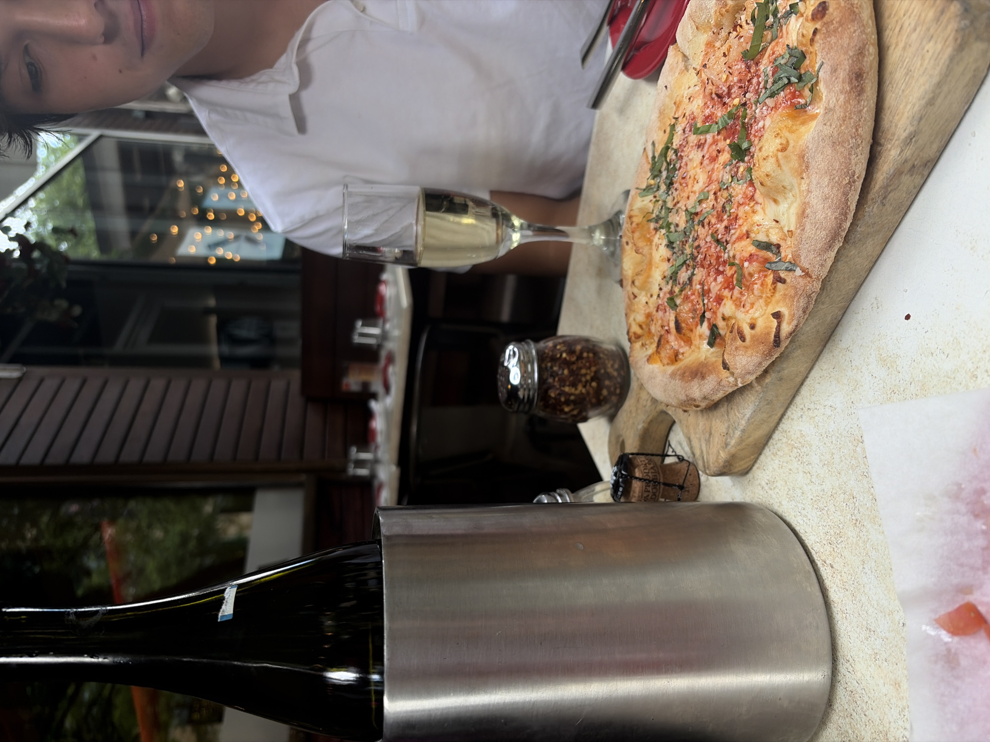 Here is a detailed description of the image:
**Scene Overview:**
The image appears to be taken in a restaurant or dining area, likely outdoors or near a window. There is a table set with a pizza, a glass of white wine, a bottle of wine in an ice bucket, a jar of red pepper flakes, and a person partially visible. The location is in the city of Austin, United States of America based on the cork.
**People:**
* A person is partially visible in the upper left corner, wearing a white shirt. Their face is visible, but only a portion of the body is in the frame.
**Subjects/Objects:**
* **Pizza:** A rectangular pizza is placed on a wooden serving board. It appears to be topped with cheese, red pepper flakes, and fresh basil.
* **Wine Glass:** A glass of white wine stands on the table.
* **Wine Bottle:** A dark glass wine bottle is encased in a metal ice bucket.
* **Red Pepper Flakes:** A glass jar filled with red pepper flakes is present.
* **Cork:** A wine cork is placed near the ice bucket.
* **Napkin:** A white napkin with red specks is placed next to the ice bucket.
**Environment:**
* The image is taken in an area with outdoor exposure. The windows look out to trees and other objects.
* The lighting suggests it is daytime.
**Actions/Interactions:**
* The scene implies a meal is in progress, or about to begin. The presence of the wine and pizza suggests a dining experience.
* There is an implication that the wine bottle came from the ice bucket due to the nearby cork.