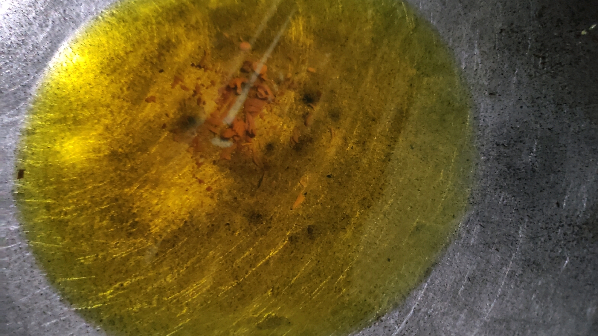 The image shows a close-up view of a shallow pool of yellow oil on a textured gray surface. There are small dark specks and some reddish-orange debris suspended within the oil, particularly concentrated in the upper central portion of the pool. The surface beneath the oil appears to be metallic and shows a pattern of fine scratches. The lighting creates reflections and highlights on the oily surface. No specific location in Notorejo, Indonesia can be determined from this image.