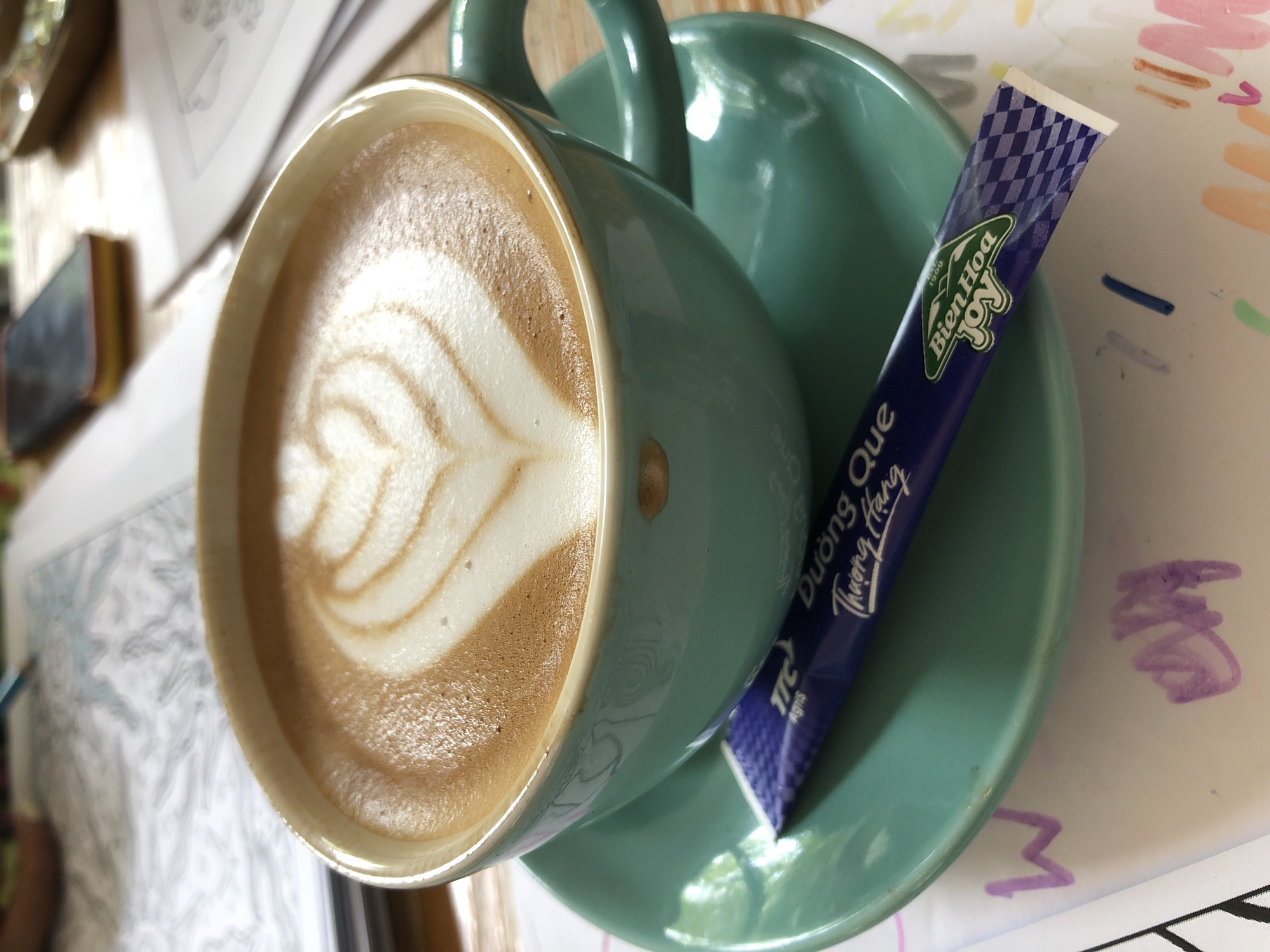 A close-up, eye-level shot shows a light blue coffee cup filled with cappuccino art, a latte art heart design, resting on a matching saucer. A purple sugar packet with Vietnamese text and a "Bien Hoa" logo lies diagonally across the saucer in front of the cup. The background is blurred but suggests a table with papers, possibly a coloring book with a pencil, and a mobile phone. This scene is consistent with a cafe setting in Đà Nẵng, Vietnam, where such artistic coffee presentations and sugar packets are common.
