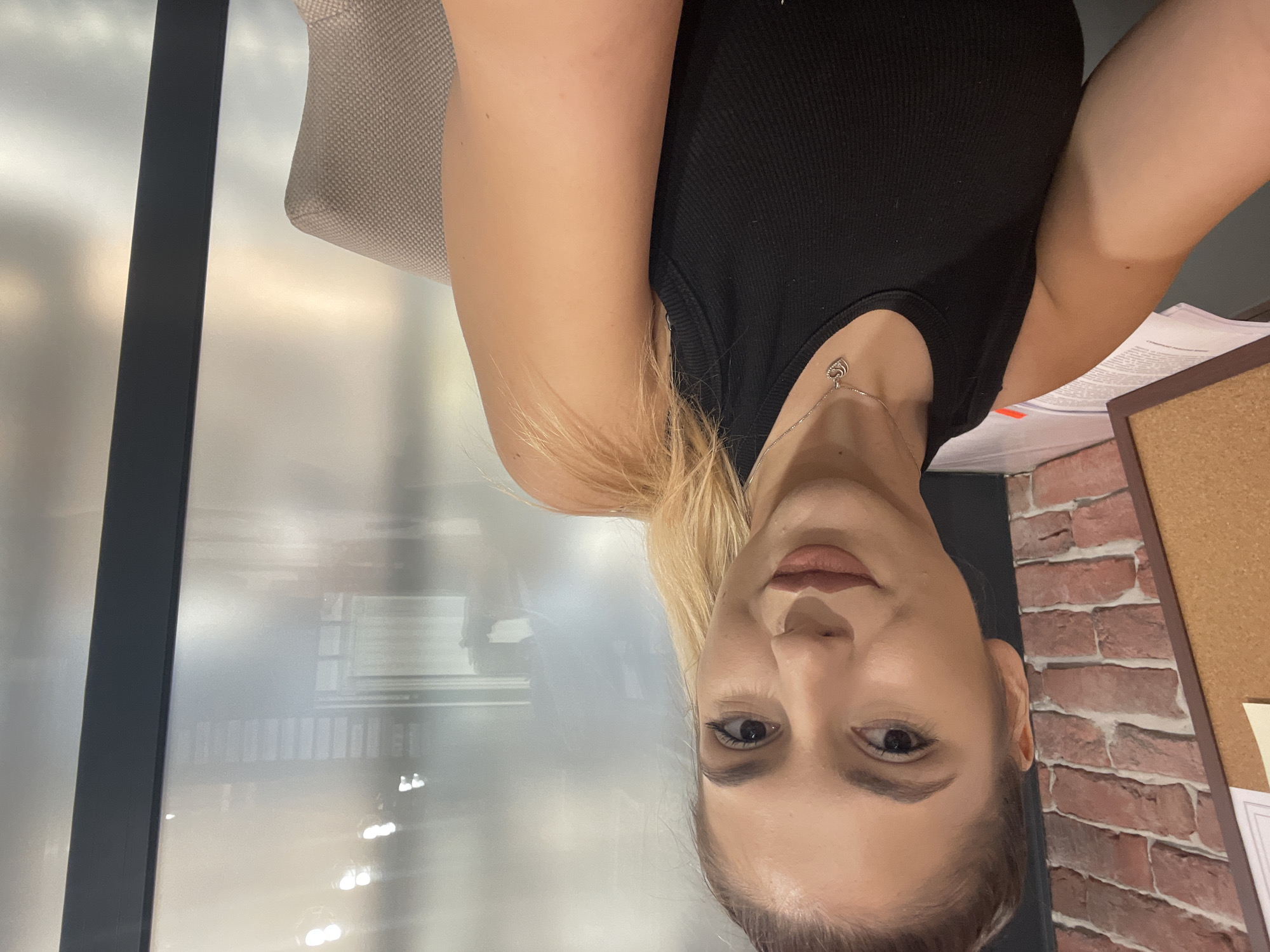 A woman with blonde hair tied in a ponytail is taking a selfie in an office environment. She is wearing a black tank top and a silver necklace with a heart-shaped pendant. The background shows a brick wall on the left, papers on a desk, and a frosted glass partition with reflections of office lighting and structures on the right. The chair she is sitting on is visible on the right side of the image. The lighting suggests an indoor setting, possibly during the daytime given the reflections. There are no specific indicators of the city of Burgas, Bulgaria within the image itself.
