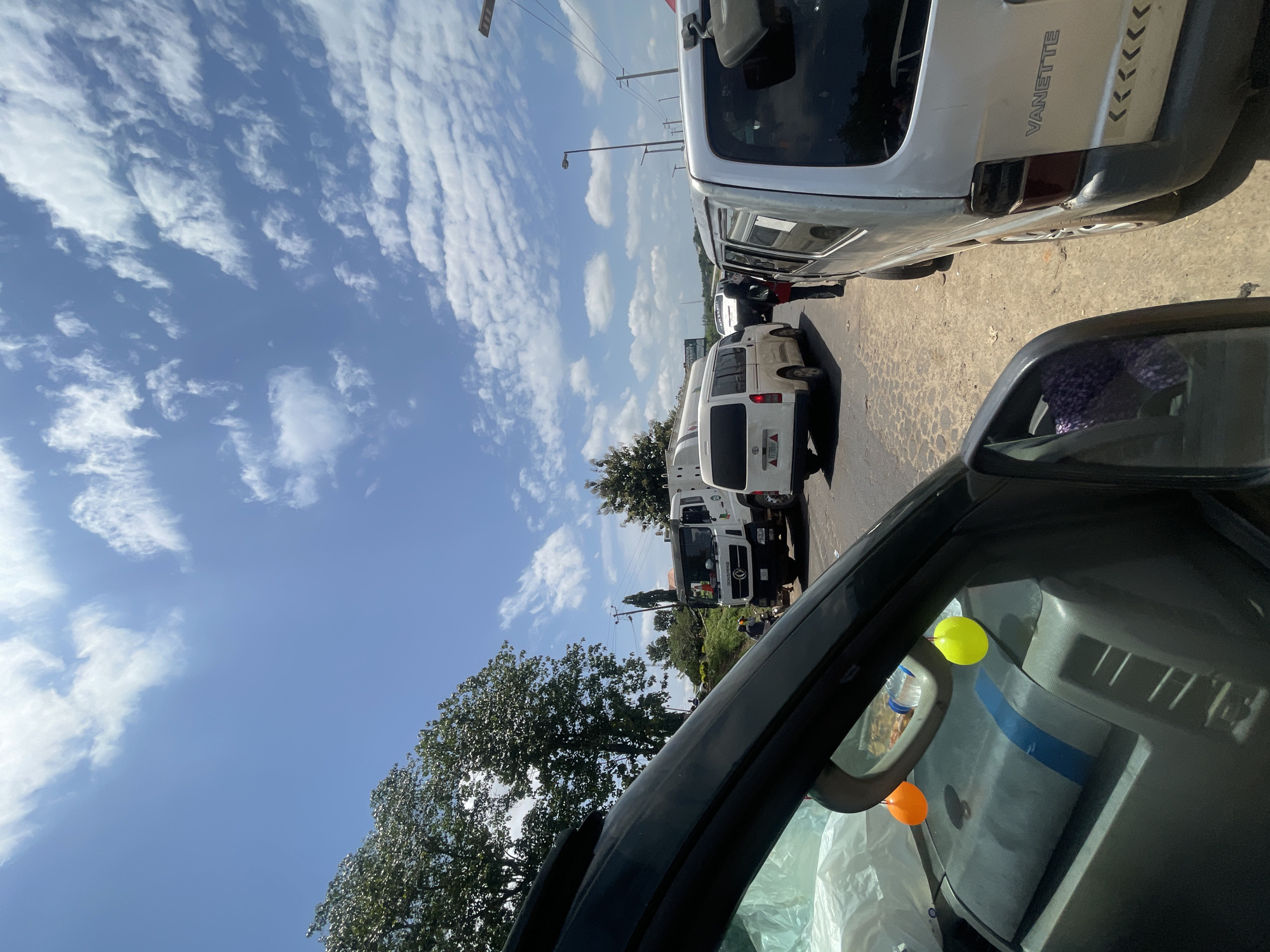 The image depicts a street scene in Gbongan, Nigeria, with several vehicles and a clear blue sky dotted with white clouds. In the foreground, the interior of a vehicle is visible, including a side mirror and part of the dashboard with two colorful hanging ornaments. Beyond this vehicle, a silver van with the word "VANETTE" on the back is parked to the right. Further down the road, a white truck with a water tank is visible, along with other vans and vehicles. There are a few individuals standing near the vehicles, appearing to be engaged in some activity. Trees line the left side of the road, and utility poles are visible in the background. The road surface appears to be paved and somewhat worn.