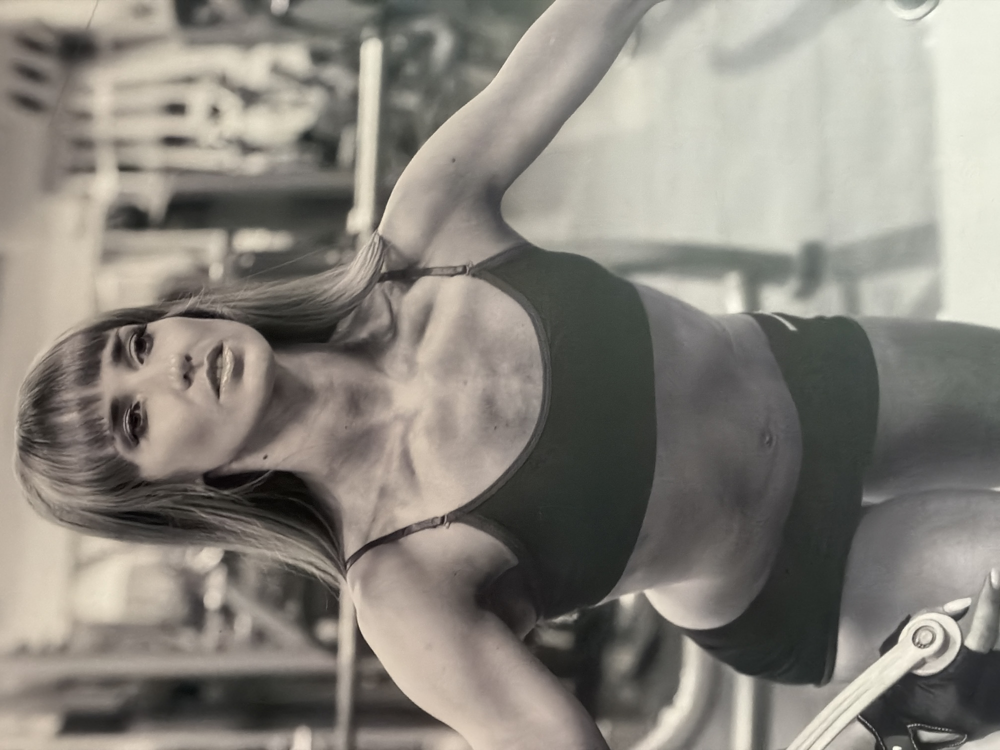 The image is a black and white photograph of a woman in workout clothes inside a gymnasium, possibly located in Izhevsk, Russia. The woman is wearing a sports bra and shorts. She has her arm raised and appears to be engaging in some kind of exercise using gym equipment. There is other gym equipment visible in the background.