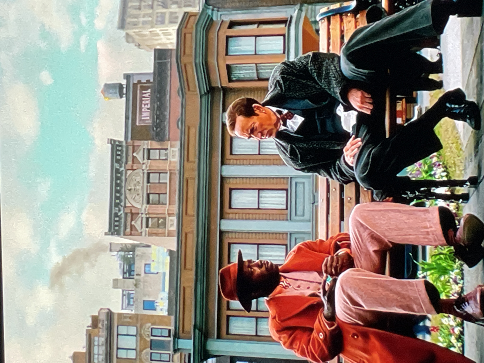 Two men sit on a park bench in front of a city scene. One man wears an orange suit with a matching hat and is in the foreground, while the other is dressed in a black suit and sits behind him. Behind the men, city buildings with windows rise into a cloudy sky. One building has the word "IMPERIAL" displayed on its side.