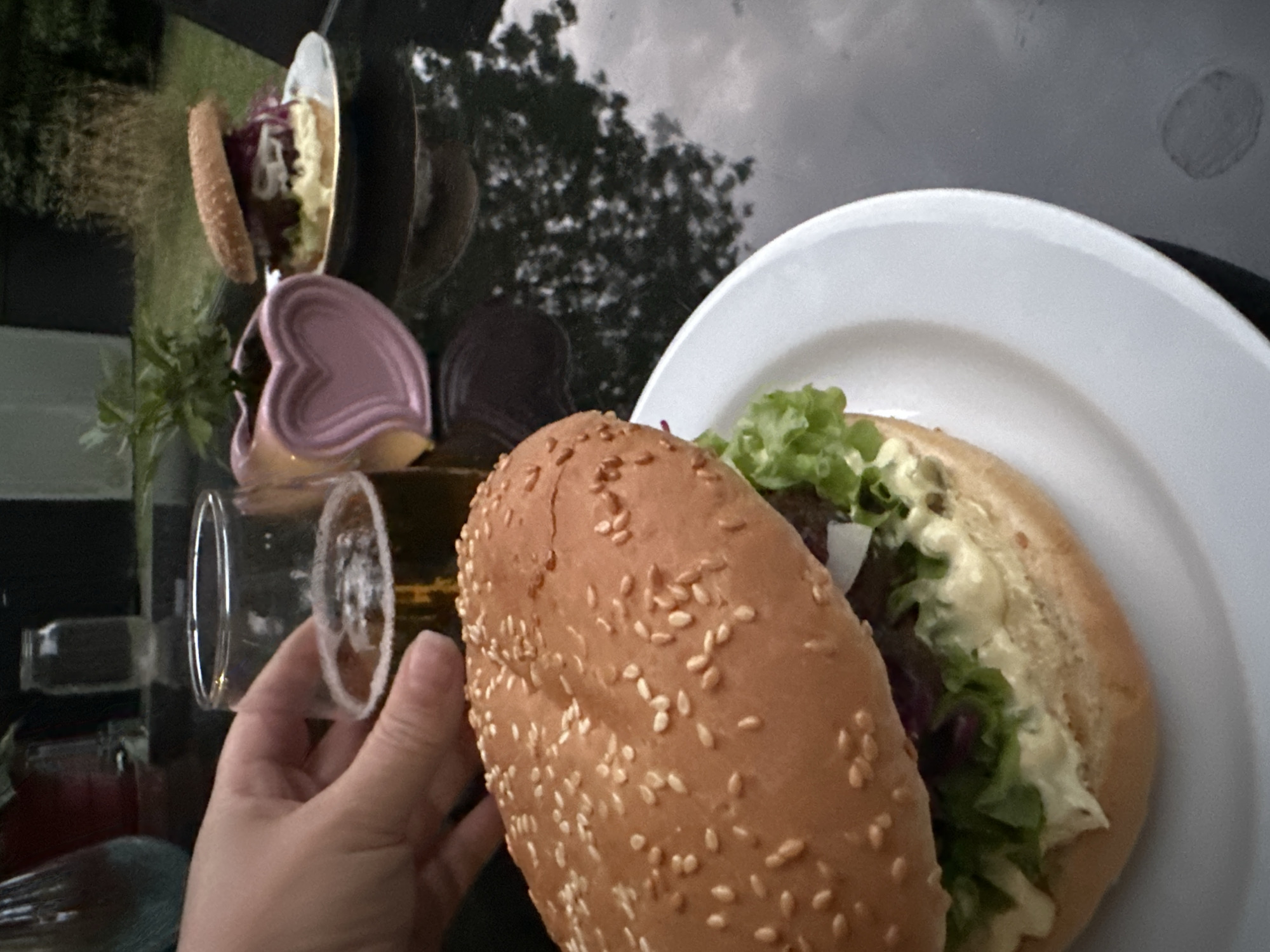 A close-up, eye-level shot captures a table setting outdoors, likely in a garden or on a patio. The foreground is dominated by a large, sesame-seed bun hamburger on a white plate. The burger is adorned with lettuce, red cabbage, and a generous dollop of mayonnaise. To the left, a hand is visible, holding a glass filled with a golden-brown beverage, possibly beer, with a frothy head.
In the background, a smaller hamburger sits on a plate, accompanied by a small pink heart-shaped planter with a green plant. The table's reflective surface shows a blurred outdoor environment with greenery and what appears to be a cloudy sky. The setting suggests a relaxed meal or gathering. The image does not provide any specific clues about the location in Krefeld, Germany.