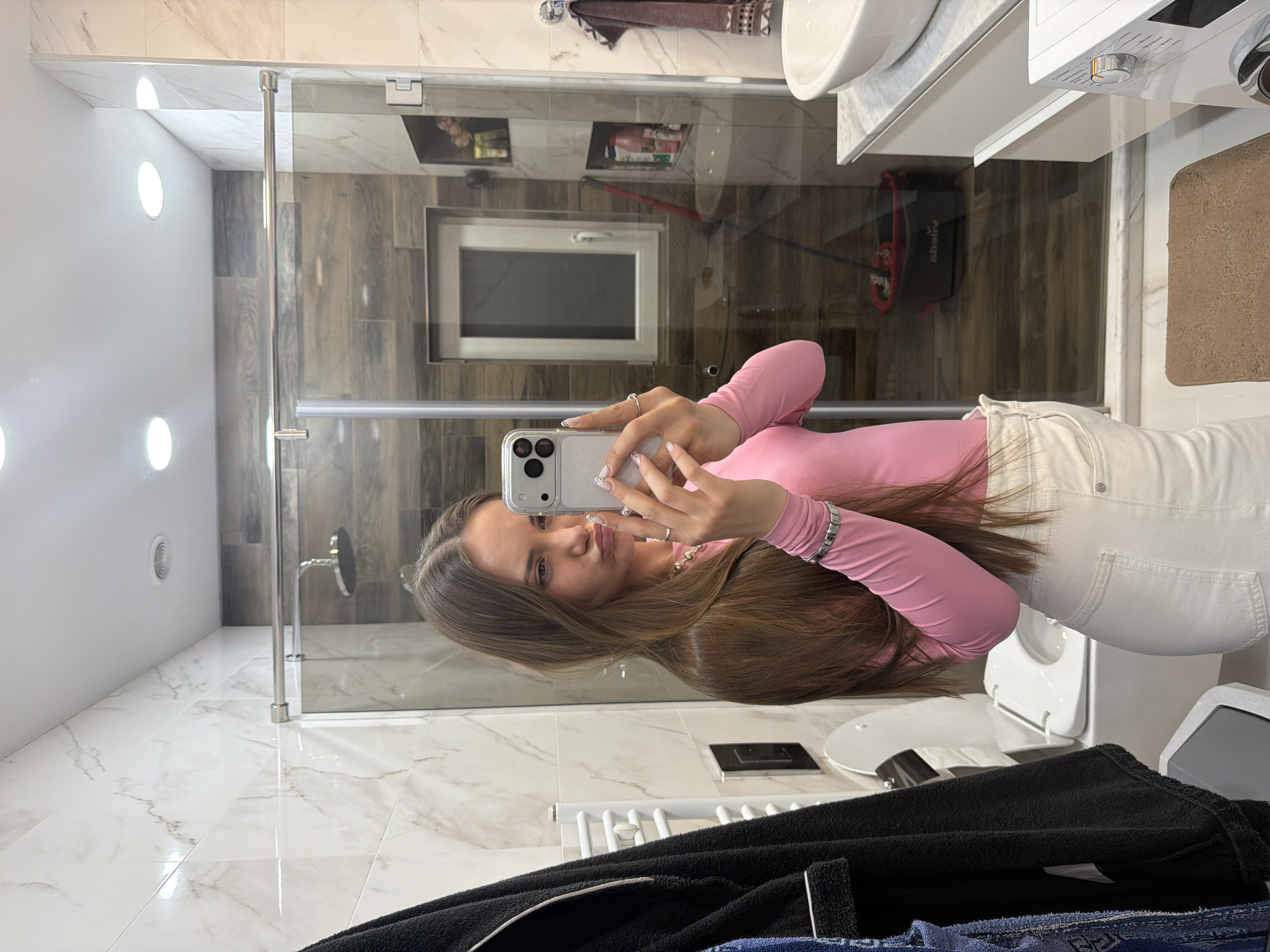 A young woman with long brown hair wearing a pink long-sleeved shirt and white jeans is taking a selfie in a bathroom. The bathroom has white marble-like tiles on the floor and wall near the shower. There is a glass shower door. The opposite wall has wood-like tiling with a window inset. A white toilet and white sink are visible in the background. A black robe and blue jeans are in the foreground. The location is likely Buhovo, Bulgaria.
