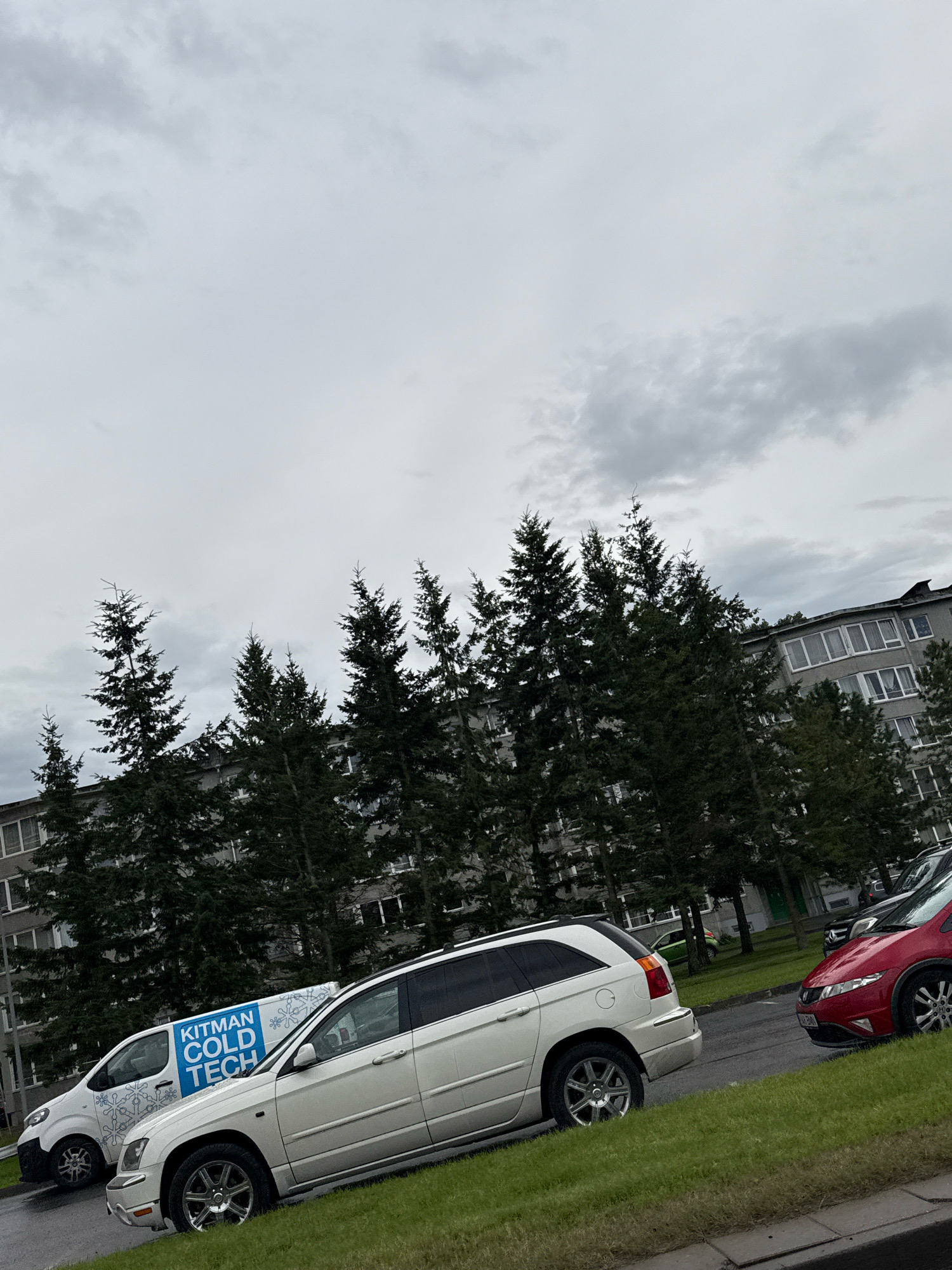 The image depicts an outdoor scene in Tallinn, Estonia, on a cloudy day. In the foreground, a white SUV is parked on the right side of the frame, facing towards the left. Next to it, to its left, is a white van with blue lettering that reads "KITMAN COLD TECH" and a snowflake design. Behind these vehicles, a row of tall evergreen trees stands prominently. Beyond the trees, a multi-story apartment building with numerous windows is visible. The sky is overcast with gray clouds. The ground appears wet, suggesting recent rain.