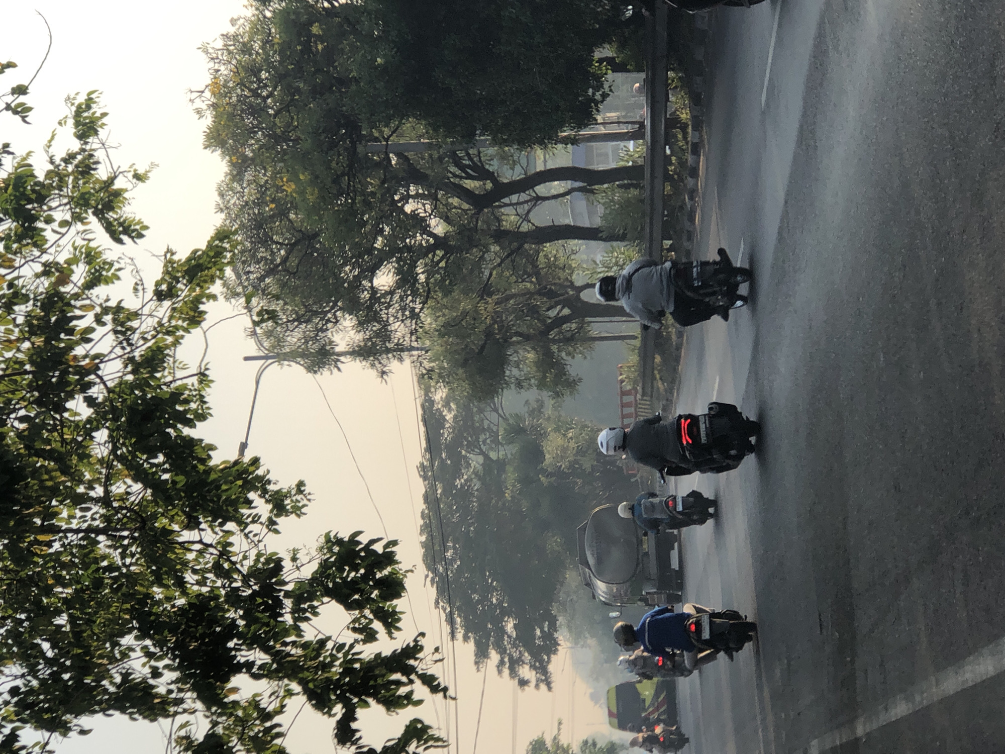 In Kludan, Indonesia, a group of motorcyclists navigates a two-lane road lined with trees and a guardrail. The road surface is asphalt, with a white lane marker visible. In the foreground, three motorcyclists are riding away from the camera. The leftmost motorcyclist is wearing a blue shirt and a white helmet. The middle motorcyclist wears a blue jacket and a white helmet, with a backpack on their back. The rightmost motorcyclist is wearing a gray jacket and a white helmet, with a black backpack. Behind them, a large tanker truck is in the right lane, also moving away from the camera. To the left of the tanker truck, a yellow and green vehicle is partially visible. The background features dense foliage, including trees and bushes, with a hazy sky suggesting early morning or late afternoon light. Electrical wires and poles are also present, indicating an urban or suburban setting.