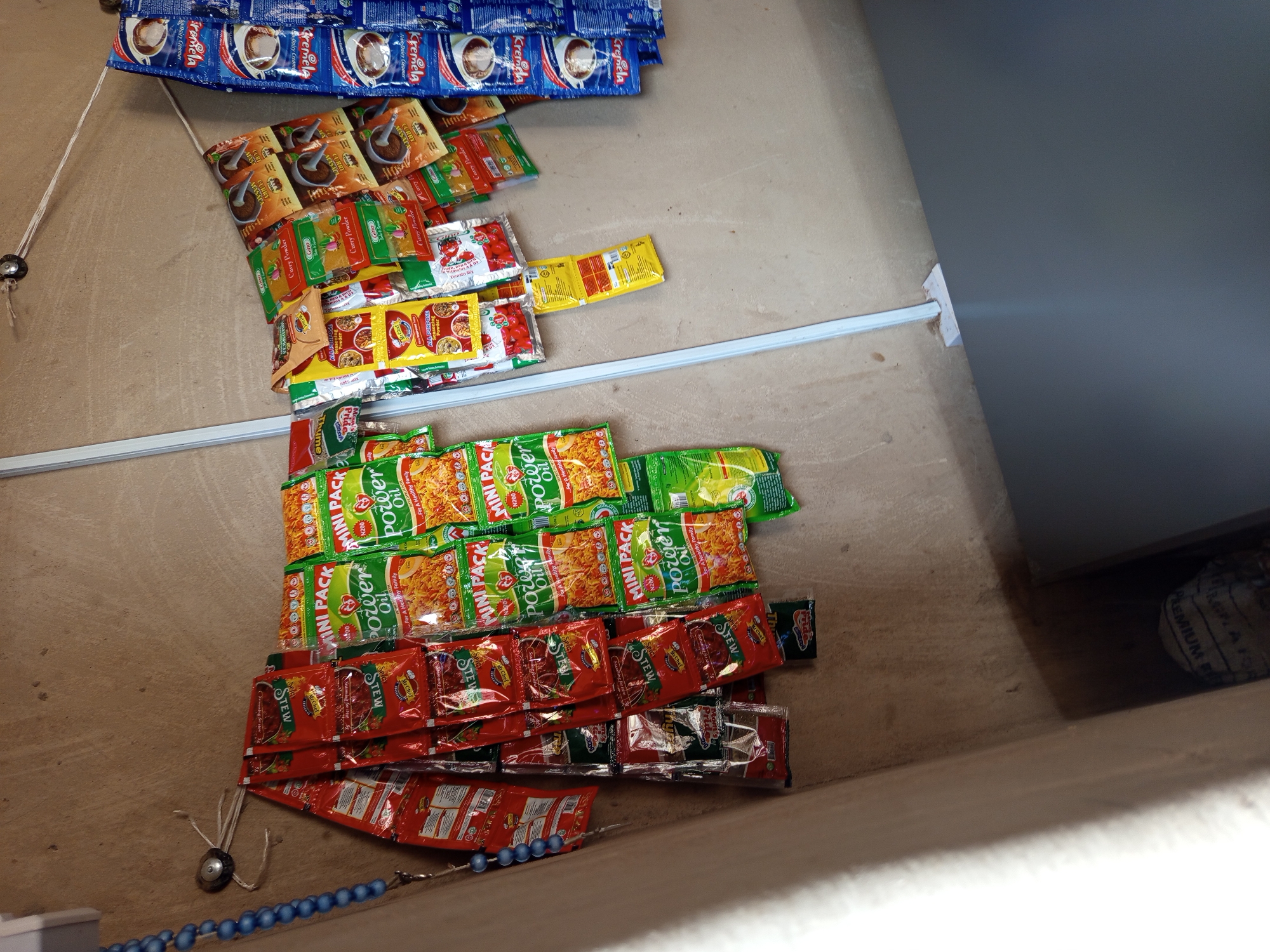 The image depicts a small retail display of various food seasoning and creamer sachets hanging from a wire against a textured wall. The sachets are colorful and feature product branding. On the left, a strand of blue beads hangs down, attached to a hook on the wall. The lighting appears to be natural, casting some shadows. The scene is likely a roadside stall or a small shop in Jalingo, Nigeria, given the context of the product packaging which often features English and sometimes local languages.
