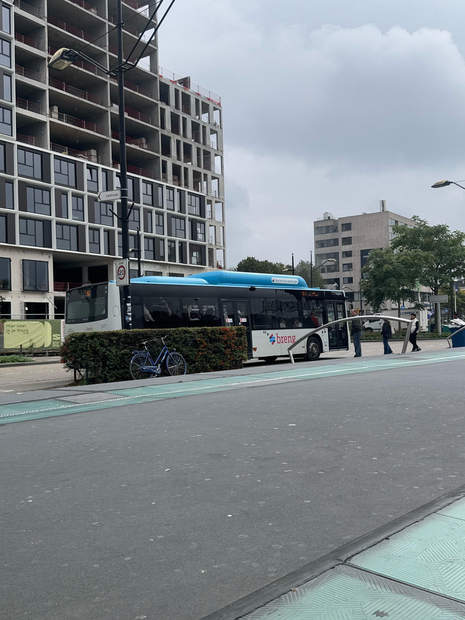 The image depicts a street scene in Nijmegen, Netherlands, likely near the city center, as indicated by a sign reading "Centrum." A modern, under-construction building with a grid-like facade dominates the background on the left. In the middle ground, a white and blue Breng bus is stopped, with its doors open. Several passengers are visible inside the bus, and a few individuals are standing on the sidewalk near the bus, appearing to be boarding or disembarking. A blue bicycle is parked in front of a dense hedge next to the bus. The foreground shows a road surface with a distinctive green, patterned pedestrian or bicycle lane. The sky is overcast, suggesting a cloudy day.