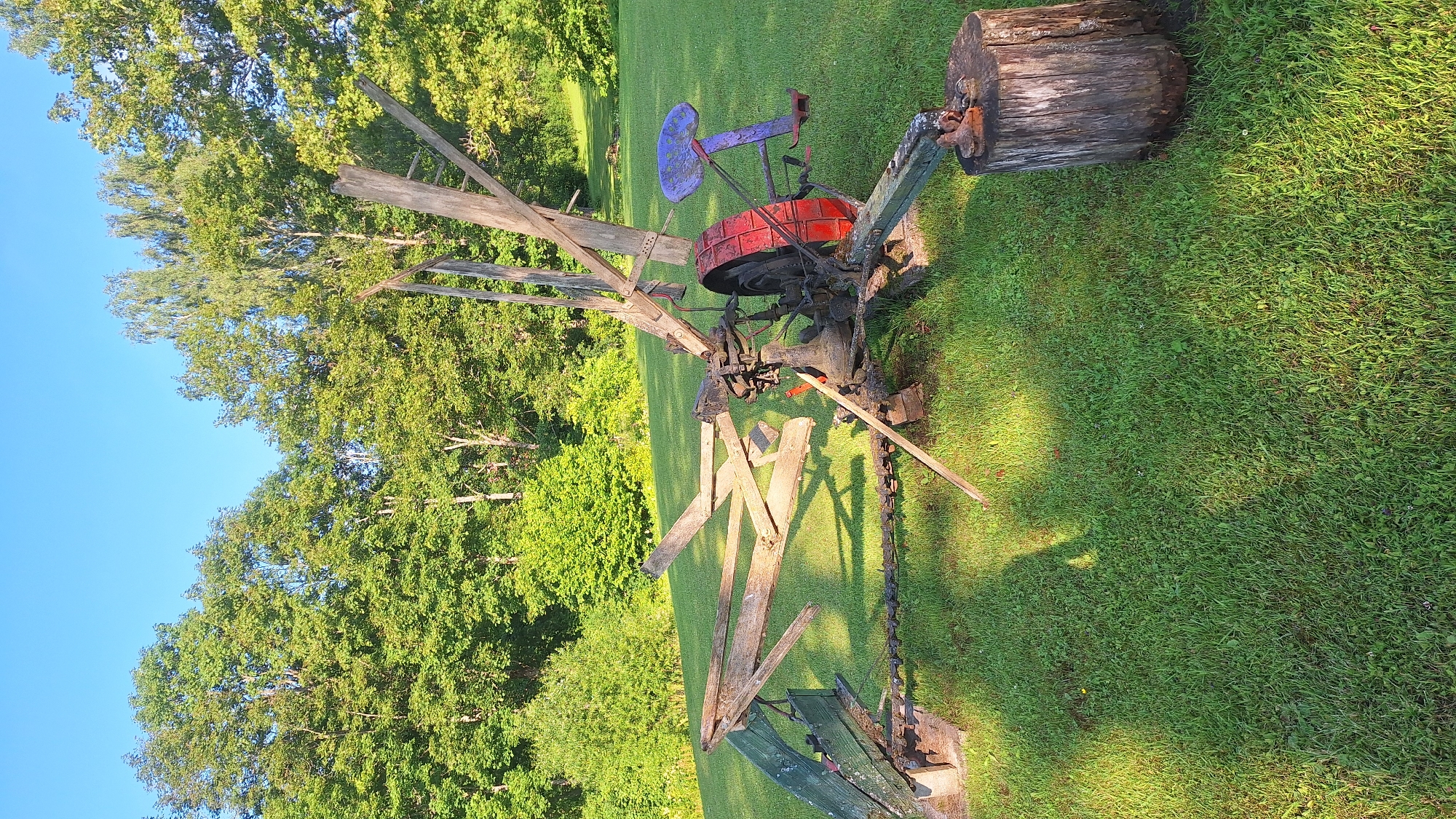 An antique agricultural machine, likely a mower or reaper, sits on a green, grassy lawn. The machine is constructed of wood and metal, with a large red wheel on one side. A purple seat is attached to the back of the machine. The machine appears to be in a state of disrepair. In the background, a line of green trees borders the lawn, and a clear blue sky is visible above. The scene is bathed in sunlight, casting shadows on the grass. The image was taken in Lēdmane, Latvia.