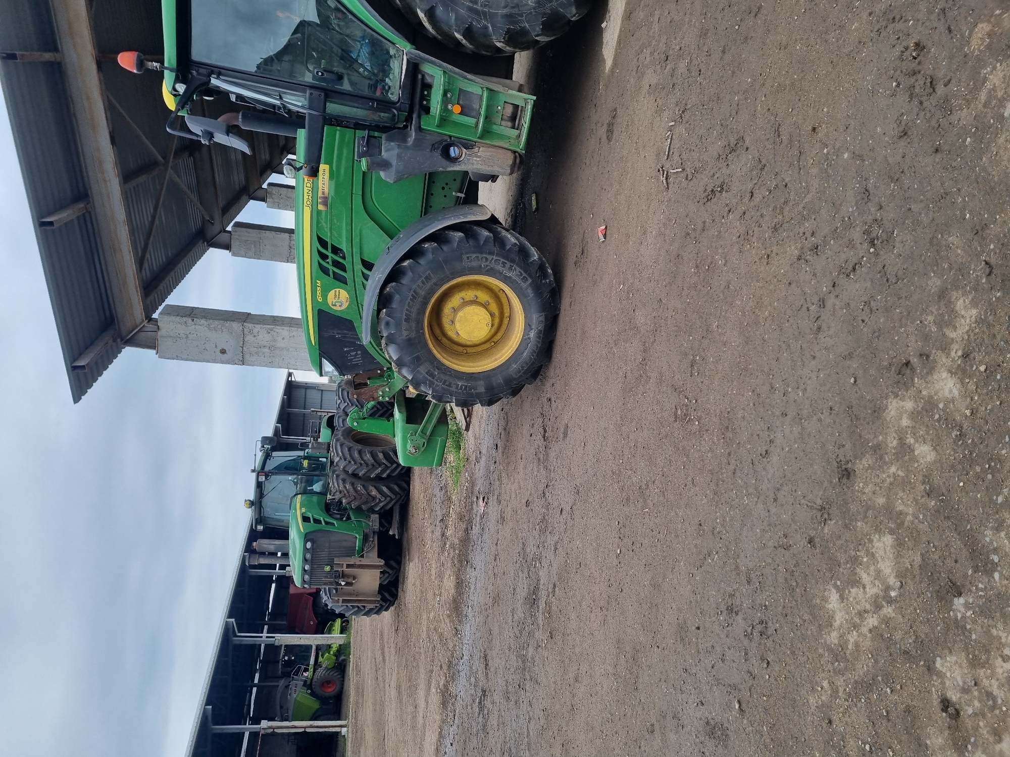 The image displays a row of large agricultural tractors parked under a substantial open-sided shelter in Ovcharovo, Bulgaria. The main focus is on a green John Deere tractor in the foreground, easily identifiable by its prominent green body with yellow detailing on the wheel hubs. Visible text on this tractor includes "JOHN DEERE," the model number "6155M," and "МЕГАТРОН" (MEGATRON), along with a "5" logo. The large front tire shows "MICHELIN" and the size "560/65R-28."
Further along the row, several other green tractors are parked, including one with a visible red wheel, suggesting it might be a CLAAS model. The shelter under which the machinery is stored features a corrugated metal roof supported by sturdy concrete pillars.
The ground beneath and around the tractors is a dirt and gravel surface, appearing damp or muddy in areas. The sky is overcast, indicating a cloudy day. No people are present, and the tractors appear to be idle or stored, awaiting their next use or maintenance. The scene depicts a typical agricultural storage or service area for farm equipment.