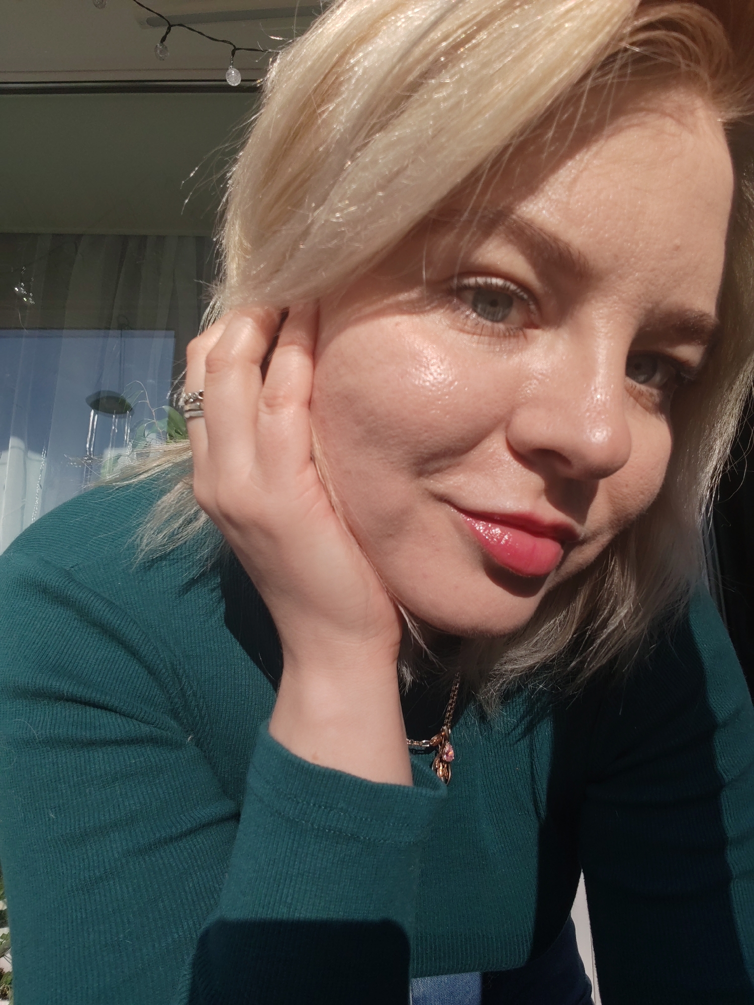 The image shows a close-up of a fair-skinned woman with blonde hair, wearing a green long-sleeved shirt and a delicate gold necklace with a pink pendant. She is holding her head with her hand, on which a ring is visible. Her lips have a shiny pink gloss, and the sunlight reflects off her skin.
The background shows a window reflecting outdoor light, with what appears to be a string of lights hanging above. There are no explicit elements in the image that would confirm a location in Izhevsk, Russia.