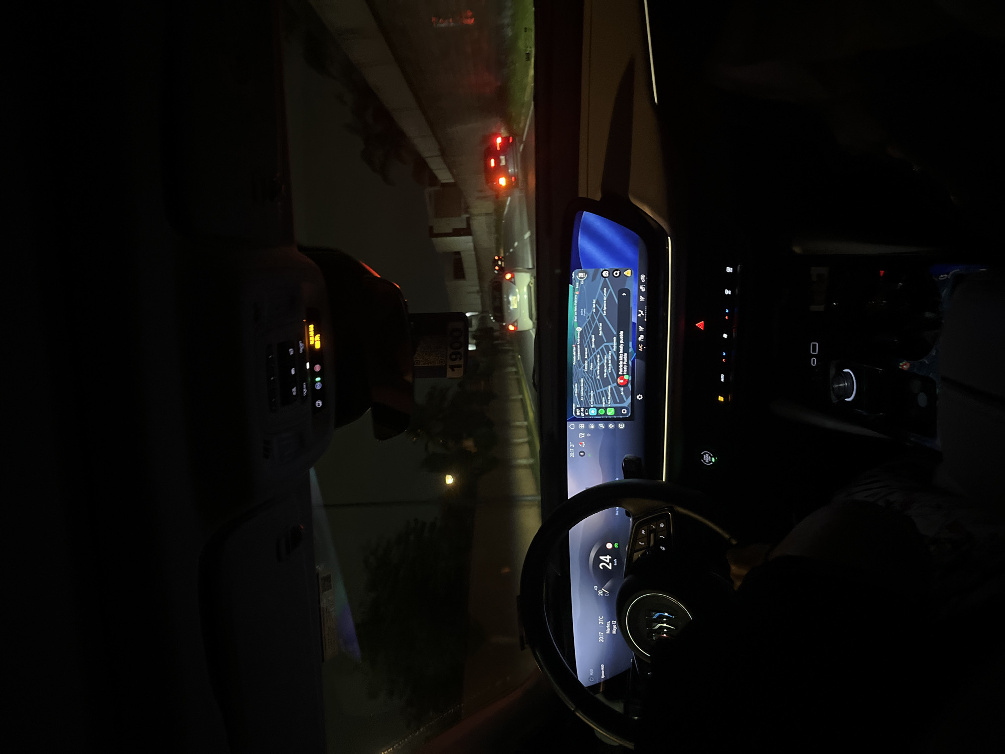 Captured from within a vehicle at night, the image presents a driver's perspective of the road ahead. The steering wheel, illuminated with digital displays indicating a speed of 24 km/h and a temperature of 21°C, is prominently featured. The dashboard integrates a large digital display, showcasing a map. The location identified through the map's OCR reads "Patricia Mtz healy puebla, Gasly Puebla, San Ignatio de Loyola" likely placing the scene near San Bernardino Tlaxcalancingo, Mexico. Outside the vehicle, there is the street with some cars ahead, the road is illuminated by car lights, streetlights, and the vehicle's headlights. The environment appears urban, suggested by the presence of buildings and other vehicles on the road.