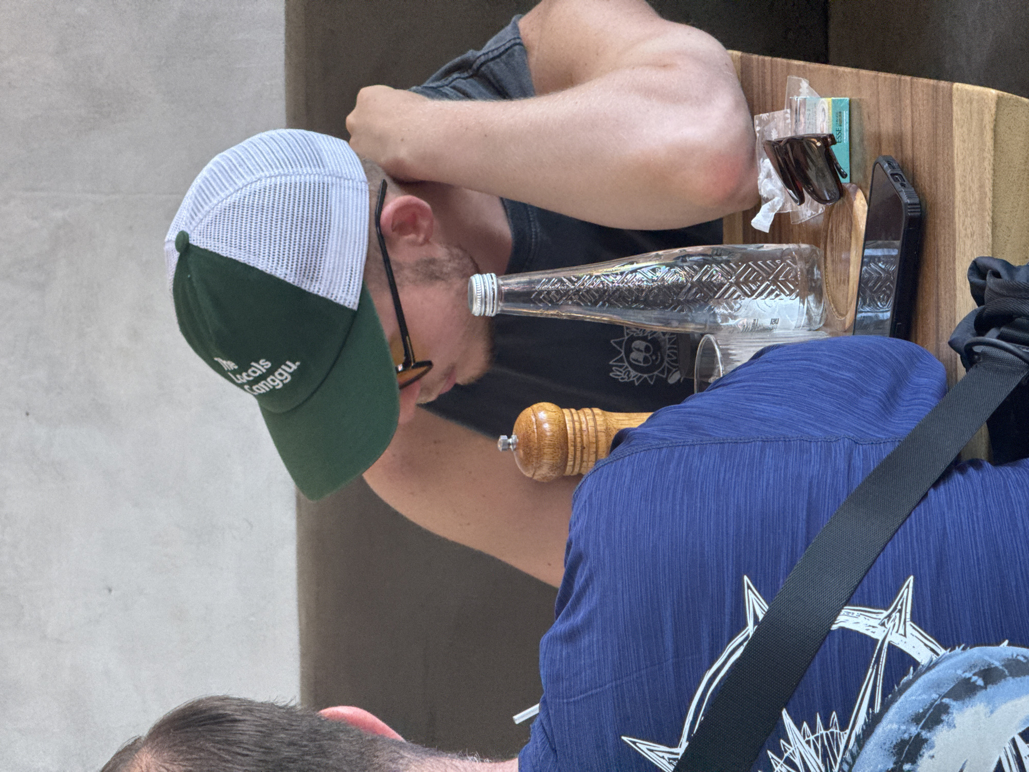The image depicts two men seated at a table, viewed from above at an angle. One man is wearing a green and white baseball cap with the words "The Locals Canggu" embroidered on the brim, suggesting a location in or near Canggu, which is near Kuta Selatan, Indonesia. He's wearing a dark gray t-shirt and glasses. His companion wears a textured blue shirt with a large white graphic design. The table holds a glass bottle, a wooden pepper grinder, a cell phone, a pack of cigarettes, and a pair of glasses. A black shoulder strap is visible, likely attached to a bag. The table is made of light-colored wood. The background appears to be a light gray wall or surface.