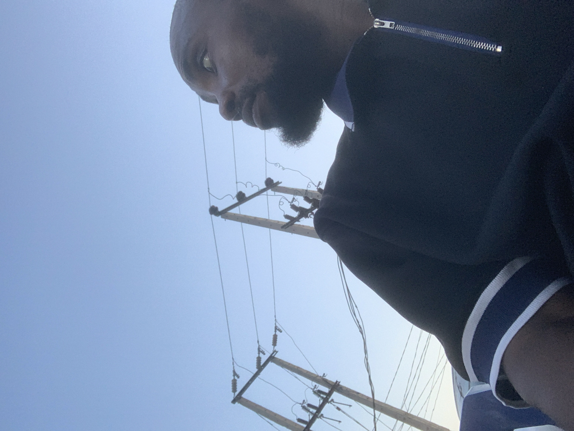 A man with a beard is positioned in the foreground, looking slightly downward. He is wearing a dark-colored top with a zipper and white trim on the sleeves. In the background, there are utility poles and wires against a light blue sky. The location is likely Sabojibi, Nigeria.