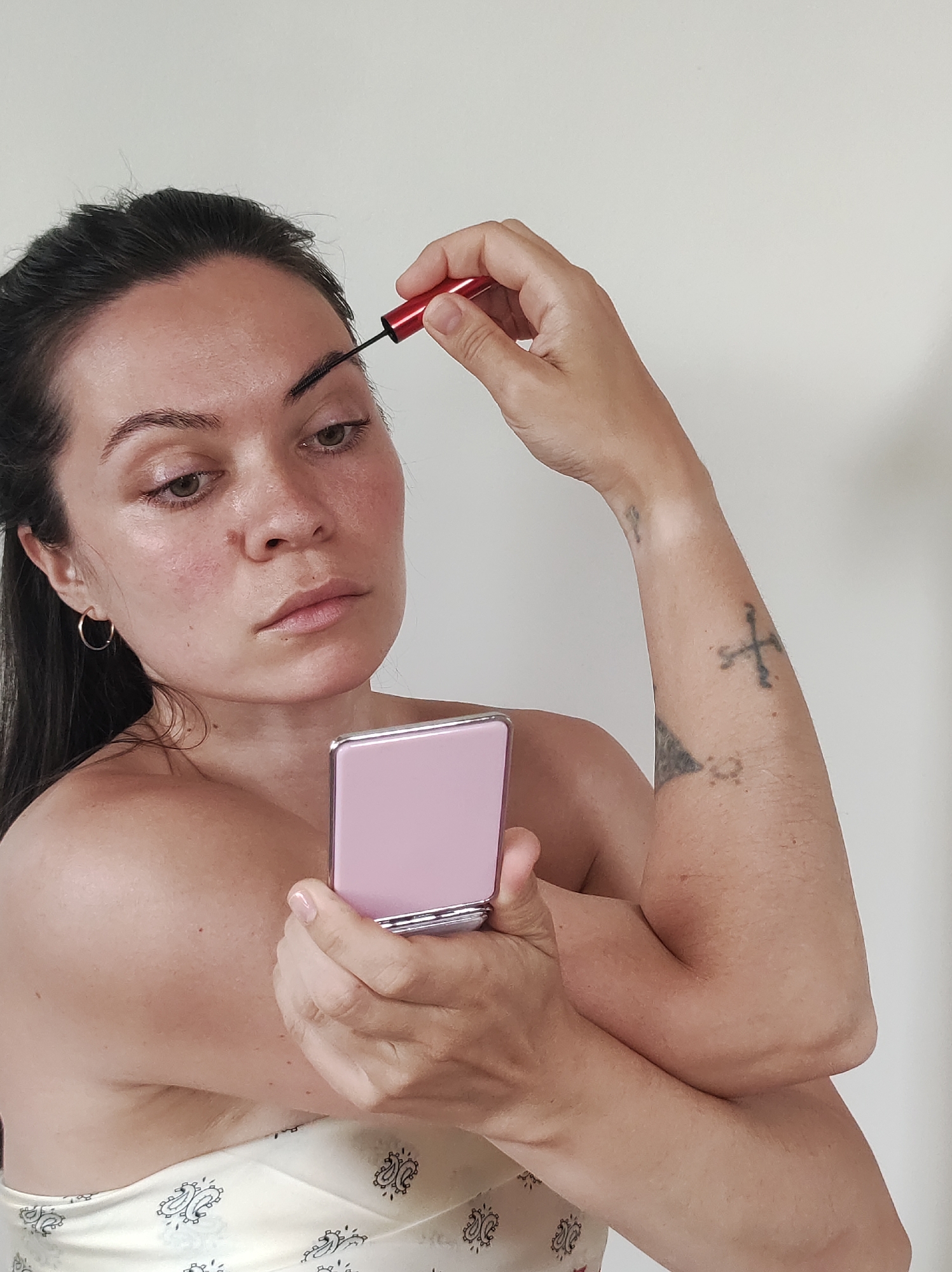 A woman is applying makeup, specifically eyebrow mascara. She is fair-skinned with dark hair pulled back. She is wearing a light-colored top with a paisley pattern. She is using a red-handled mascara wand and a pink compact mirror. Her left arm has tattoos. The background is a plain white wall.
The location in the city of Фоки, Russia cannot be confirmed from this image.