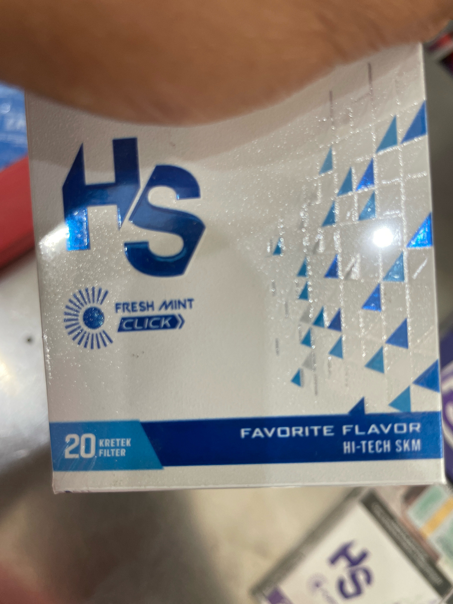 An out-of-focus, eye-level medium shot shows the side of a white and blue cigarette pack. The pack is adorned with a geometric pattern of blue and silver triangles. Prominently displayed on the pack is the brand name "HS" in large, bold blue lettering. To the left of the brand name, there is a circular graphic resembling a sunburst or stylized flower, with the text "FRESH MINT" and "CLICK" below it. The bottom of the pack features a blue band with the text "20 KRETEK FILTER" on the left and "FAVORITE FLAVOR HI-TECH SKM" on the right. The background is blurred, with hints of a hand holding the pack and other merchandise.