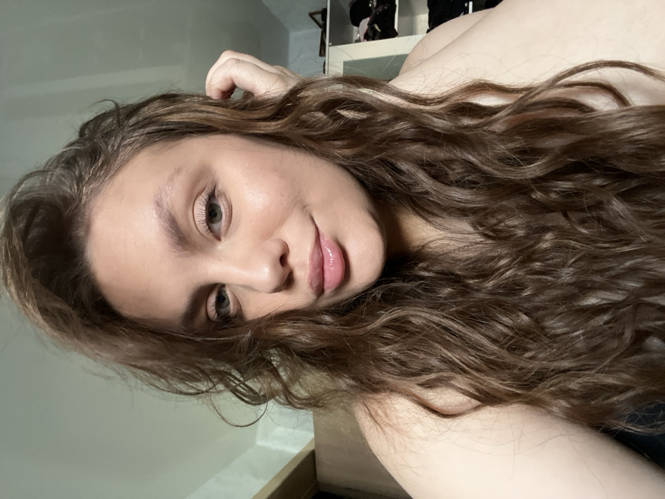 The image presents a close-up portrait of an adult female, facing forward with a slight tilt of her head. She possesses long, wavy brown hair that frames her face and extends over her shoulders. Her eyes are light-colored, and her lips are full and glossy, showing a subtle, closed-mouth smile. Her skin tone is fair. Her left hand is raised, with fingers gently touching her hair near her left ear. Her right shoulder is bare, indicating a strapless garment or bare skin. The background is out of focus, revealing elements of an indoor setting. To the left, a vertical strip of light-colored wooden trim, possibly from a door frame or wall, is partially visible. To the right, a white vertical shelving unit or closet is discernable, with dark items stored on its shelves. The lighting is soft and even. The image lacks specific visual cues to identify a precise location within the city of Chaikovsky, Russia; the environment appears to be a generic residential interior.