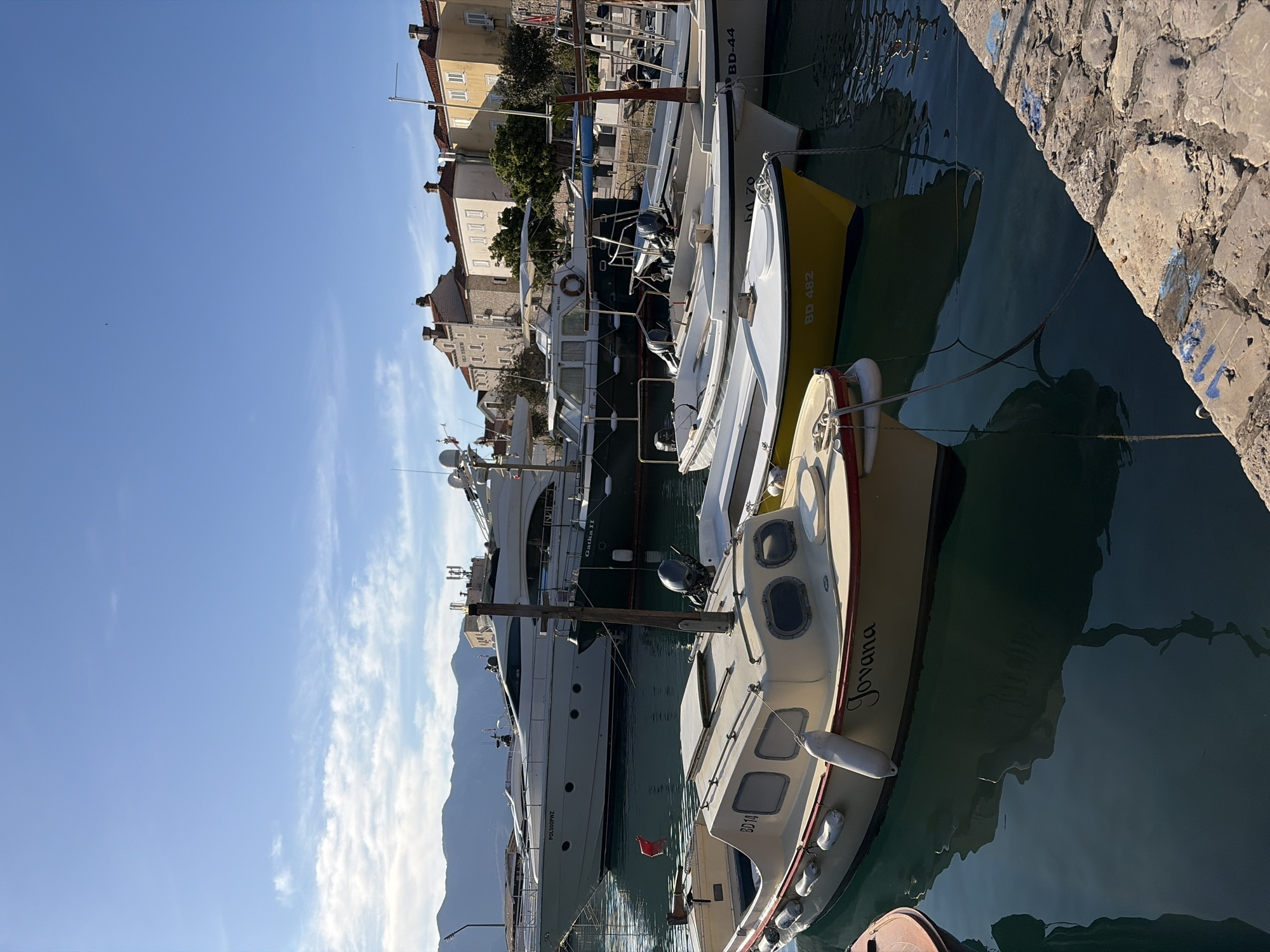The image captures a port scene in Budva, Montenegro, where several boats of different sizes are moored along the pier. The foreground features a small, light-colored boat named "Jovana" with the license plate "BD14," anchored close to the viewer. Adjacent to it is another boat, painted in a white and yellow combination, marked with the code "BD 482." Beyond these smaller vessels, a larger yacht, named "Gatka II" is docked, suggesting a luxury or recreational purpose. The buildings of Budva form the backdrop, with traditional architecture and varying heights. The sky is clear with minimal clouds, indicating sunny weather. The water in the port is relatively calm, reflecting the docked boats and the nearby cityscape. The pier is constructed with stone blocks.