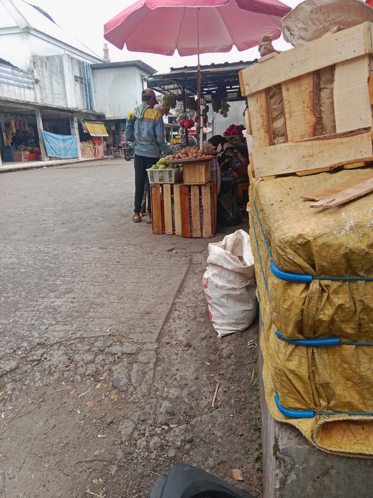 A man wearing a yellow and blue jacket and dark pants stands near a market stall in Batur, Indonesia. He is facing away from the camera, looking towards the stall which displays a variety of fruits and vegetables. The stall is covered by a large pink umbrella, and in front of it are several crates filled with produce. To the right of the stall are stacked yellow tarps covering large objects, secured with ropes and blue plastic holders. A white sack is placed on the ground next to the tarps. The ground is paved with a rough, grey material, and there are scattered stones and debris. In the background, a building with a white facade and a blue banner is visible.