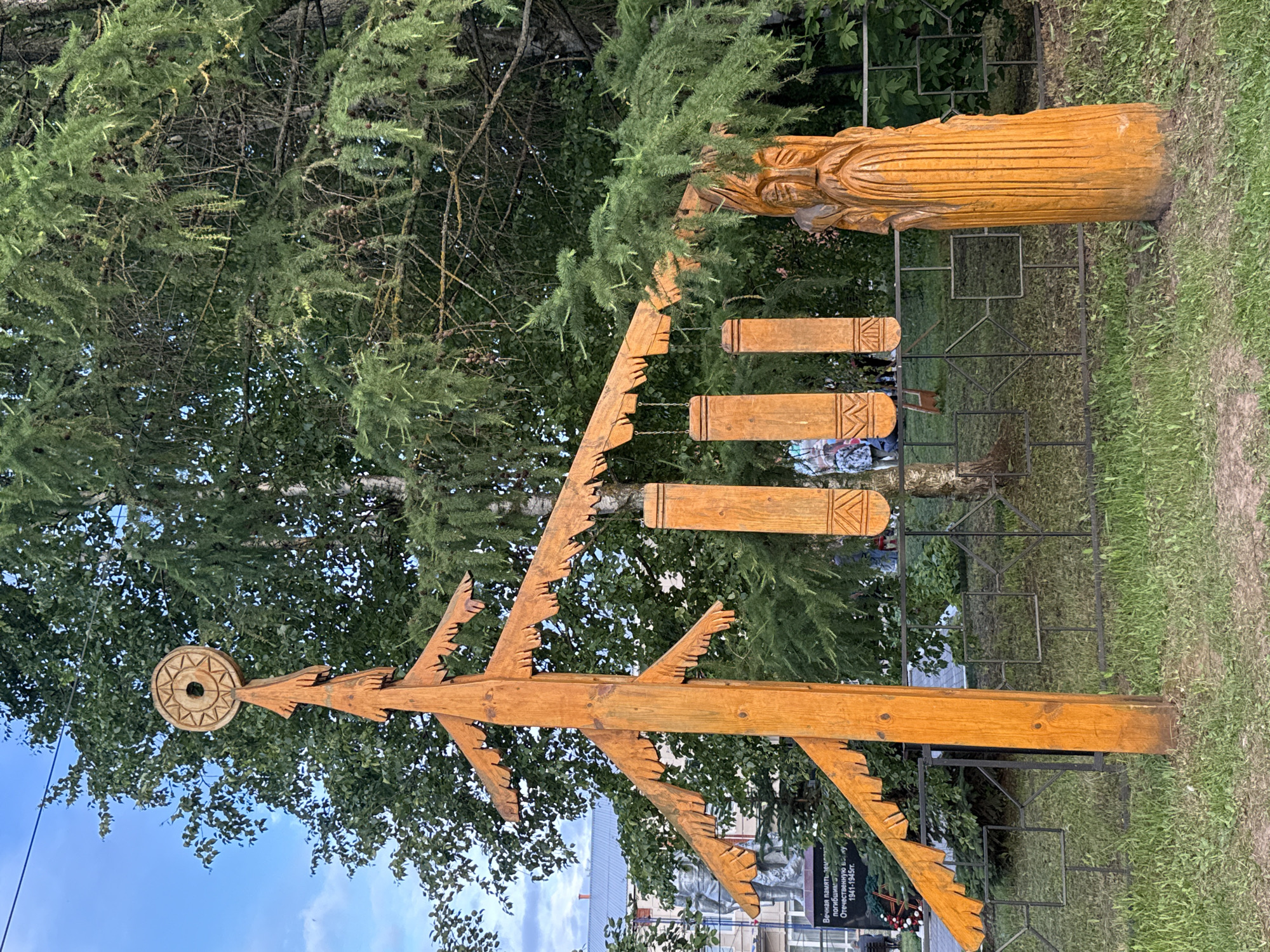 In the city of Kukui, Russia, an outdoor scene features a wooden sculpture resembling a stylized fir tree, painted orange. The sculpture has horizontal branches with jagged edges and a circular, sun-like ornament at its apex. Three rectangular wooden pieces, also painted orange and carved with geometric patterns, hang from a horizontal arm of the tree sculpture by chains. To the right of the tree sculpture is another wooden carving of a face with a beard, also orange. A dark metal fence with diamond-shaped openings runs horizontally across the middle ground. Behind the fence, on the left, a monument with a statue and an inscription is visible. The environment includes green grass in the foreground, trees and foliage in the background, and a cloudy blue sky above. A few individuals are visible in the distance behind the fence.