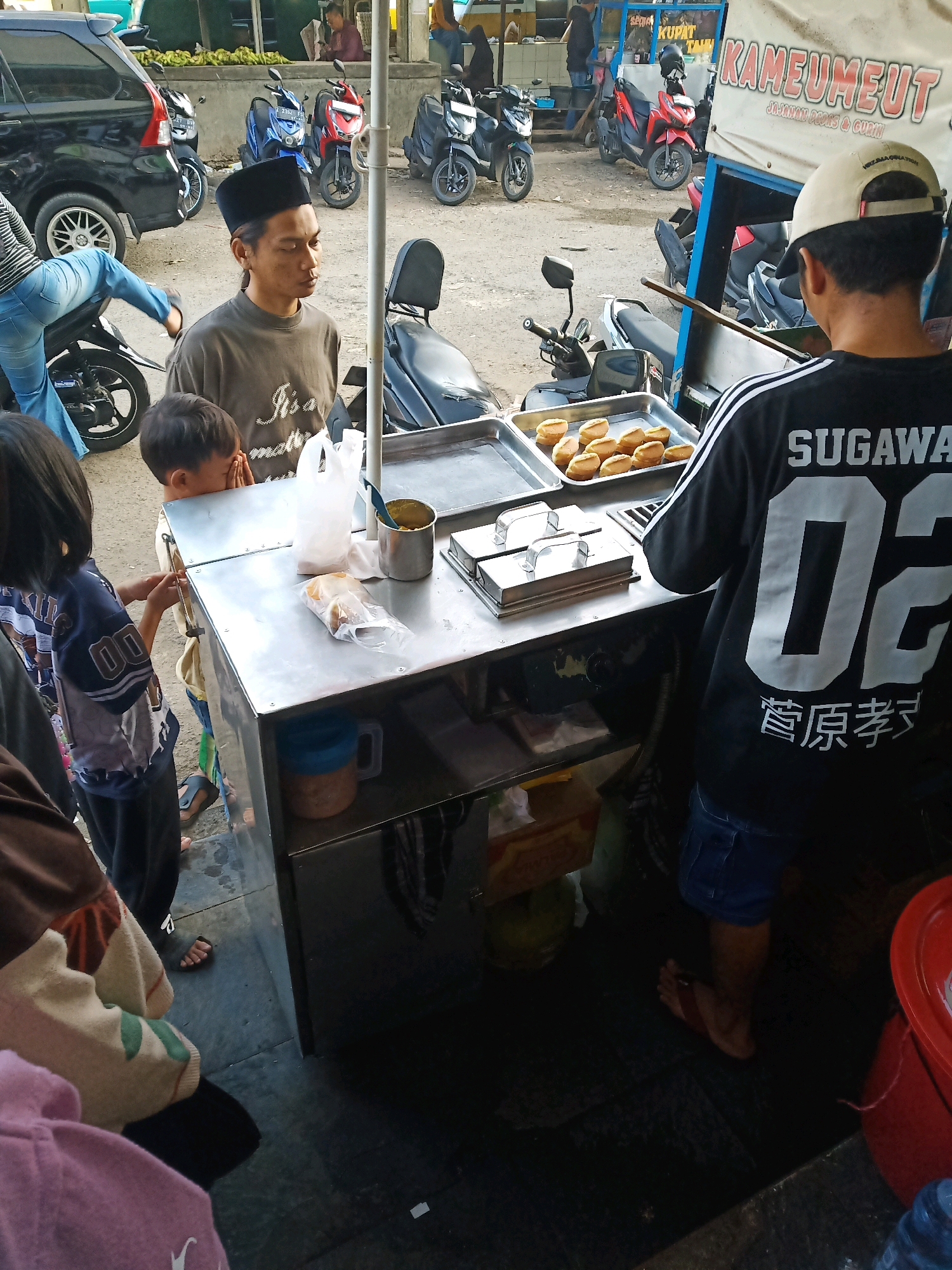 The image depicts a street food vendor in Garut, Indonesia, preparing and selling "Kameumeut," a type of jajanan (snack). A man wearing a peci (traditional cap) stands behind a metal cart with a display of cooked snacks. He is in the process of serving a customer. The cart is filled with trays of the prepared snacks, and there are also containers for ingredients and serving utensils. In the background, several motorcycles are parked, suggesting a busy street or market area. The overall scene is informal and dynamic, characteristic of a typical street food vending operation in Indonesia.