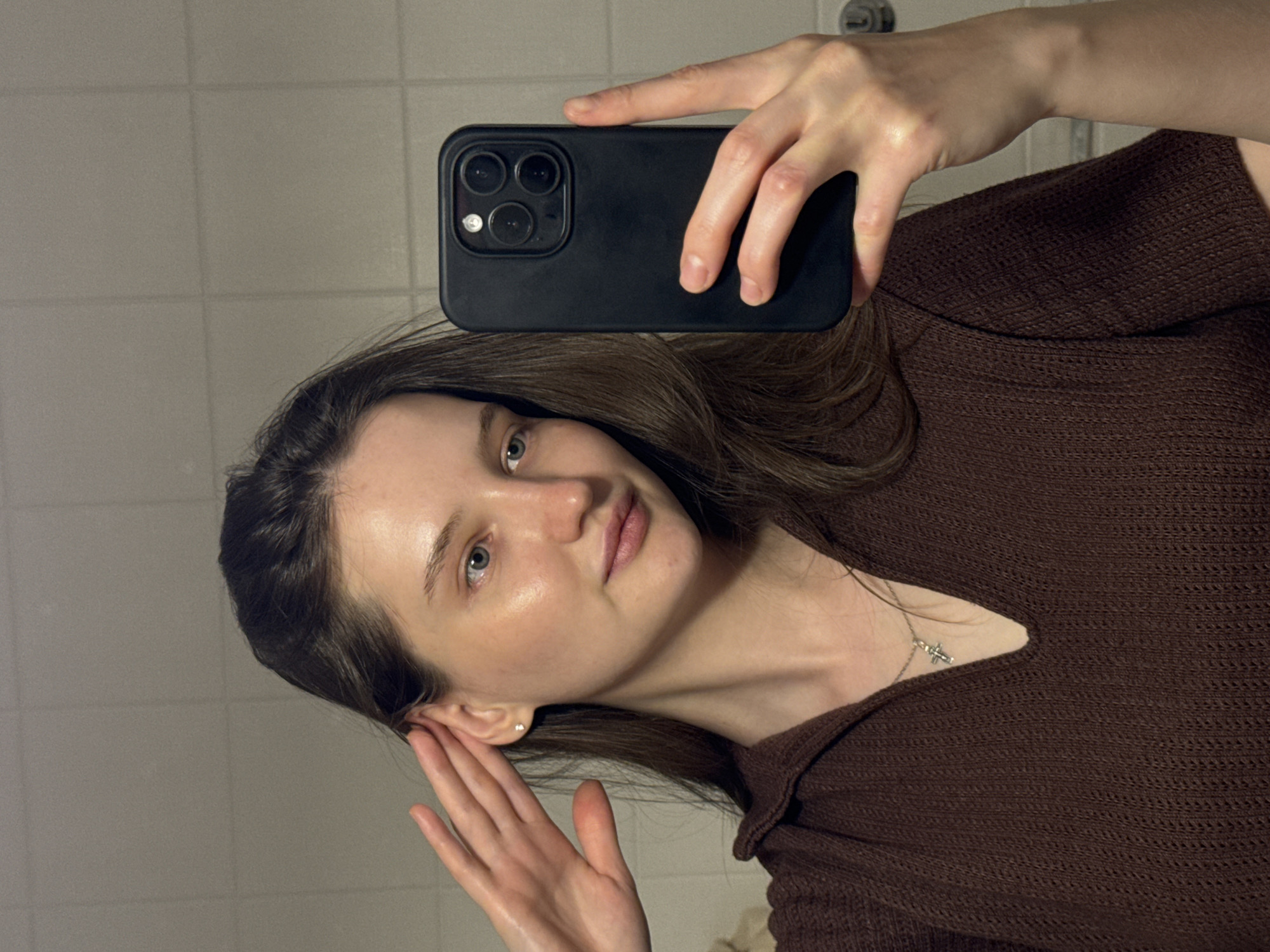 The image is a vertical, selfie-style photograph taken indoors. The primary subject is a young woman with long, dark brown hair, fair skin, and light-colored eyes. She is wearing a dark brown, textured knitted top and a delicate silver necklace with a cross pendant. Her right hand is holding a black smartphone to take the picture, and her left hand is touching her ear.
The background appears to be a tiled wall, suggesting a bathroom or a similar interior space. The lighting is soft and somewhat dim, possibly indicating evening or a poorly lit room. The woman's expression is neutral, with a slight tilt of her head. There is no visible text in the image. The overall impression is of a casual, personal snapshot. The provided location context of Журавлево (кемеровский), Russia, places the setting in a specific region of Russia, though there are no direct visual cues within the image to confirm this location.