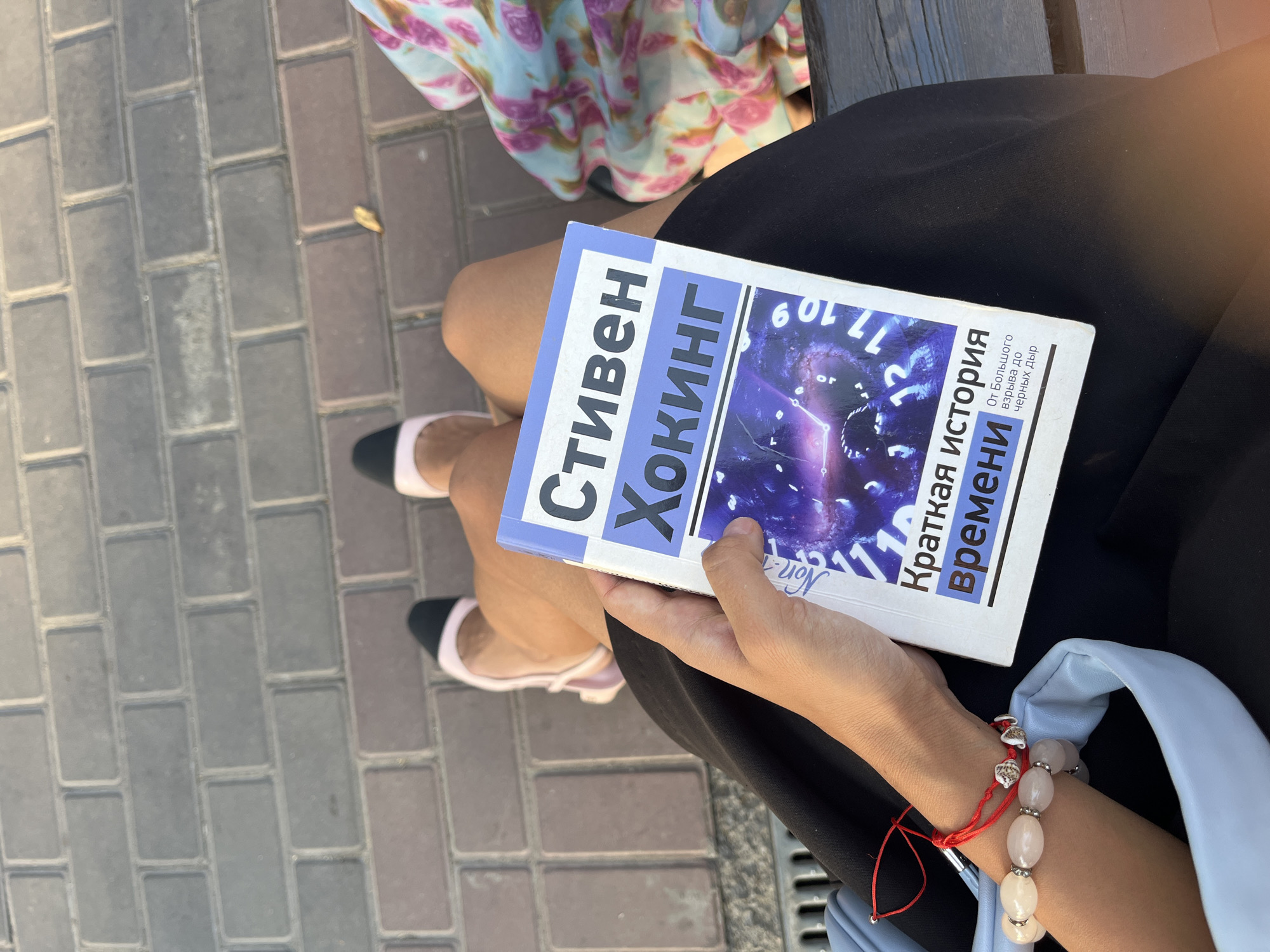 A first-person perspective shot shows a person sitting on a wooden bench, holding a book titled "A Brief History of Time" by Stephen Hawking. The book is partially open and the cover features a cosmic image with clock numerals. The person is wearing a black skirt and light blue shoes with black tips. A bracelet made of light pink beads and seashells is visible on their wrist, along with a red string bracelet. To the right, a person in a floral print dress is partially visible. The background consists of a brick pathway, suggesting an outdoor urban setting. The lighting indicates it is daytime.