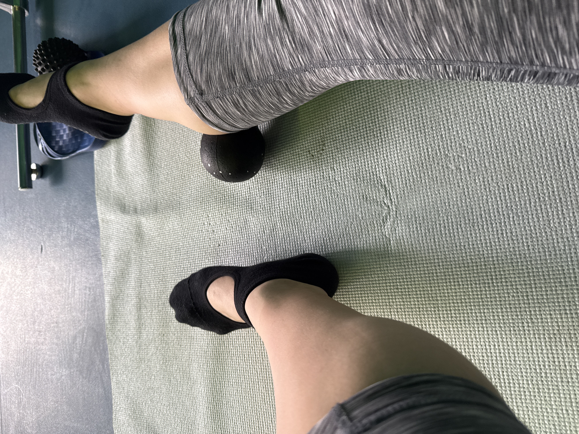 A top-down shot shows a person sitting on a pale green yoga mat, wearing gray and white striped leggings. One leg is bent with a black massage ball under the thigh, close to the knee. The person is wearing black socks. The scene is a room with a blue floor. A rack or stand with a blue container is also visible to the left. There is no information provided that indicates this image was taken in the city of Chaikovsky, Russia.
