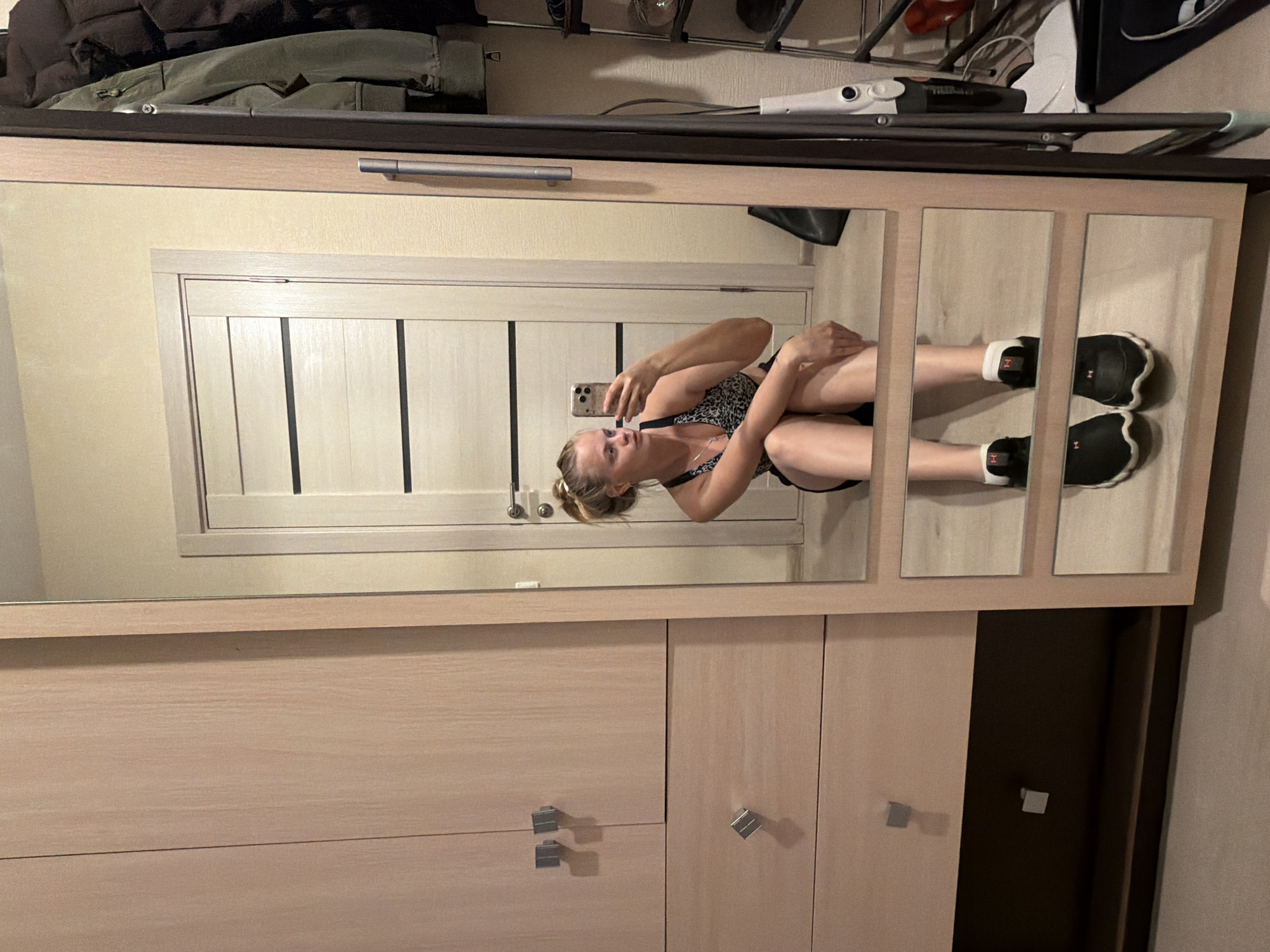 The image depicts an indoor scene featuring a young adult female taking a mirror selfie. The geographical location of Izhevsk, Russia, cannot be confirmed from the visual evidence presented in the image, which shows a typical residential interior.
The woman, positioned centrally within the mirror's reflection, is seated on a light-colored wooden floor. She has blonde hair tied back in a bun and is wearing a patterned (likely leopard print) sports bra and dark shorts. Her feet are clad in black athletic shoes with white soles and white ankle socks, each featuring an orange "H" logo. She holds a light-colored smartphone with multiple rear cameras in her right hand, aiming it towards the mirror. Her expression is neutral, and her gaze is directed at her reflection.
The mirror, framed in light wood, is a full-length piece, divided into three horizontal reflective sections by thin wooden crossbars. This mirror appears to be integrated into a larger light wood furniture unit, possibly a wardrobe or cabinet, which has a vertical silver handle on its right side. To the left of the mirror, a separate light wood wardrobe or dresser unit is visible, featuring two upper doors and one central drawer, all equipped with square, silver-toned handles. A darker brown section forms the base of this unit.
Reflected in the mirror, to the left of the woman, is a light-colored interior door with three horizontal dark panels and a silver handle. Behind her, a dark bag rests on the floor. To the right of the woman's reflection, a dark metal clothes rack holds several garments, including a dark green jacket and a dark puffer jacket. Next to the clothes rack stands a white and dark-colored stick vacuum cleaner. Further right, a small, dark-colored tiered shelving unit is visible, displaying a few small objects. The walls are painted a light beige or cream color. The overall environment suggests a well-maintained, functional living space.