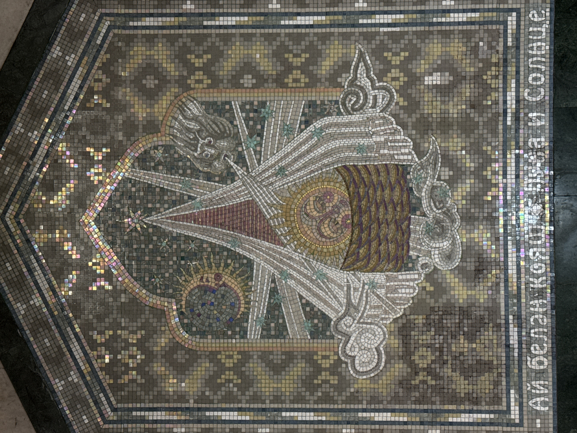The image shows a mosaic tile floor in the Kazan Metro, specifically at the Kremlyovskaya station in Kazan, Russia. The mosaic depicts a stylized emblem or symbol, featuring a central circular motif with radiating lines resembling rays or spears. The colors of the mosaic vary from earth tones and muted golds to whites and reds. Around the central symbol, there are representations of what appear to be stars, a sun, and decorative elements with flowing or flame-like shapes. The background of the mosaic is made up of small, square tiles in a repeating pattern of brown and gold. Along the right side of the mosaic are Cyrillic characters forming text. The image is taken from a slightly angled, elevated perspective, showing the mosaic in its physical context as part of a larger floor surface.