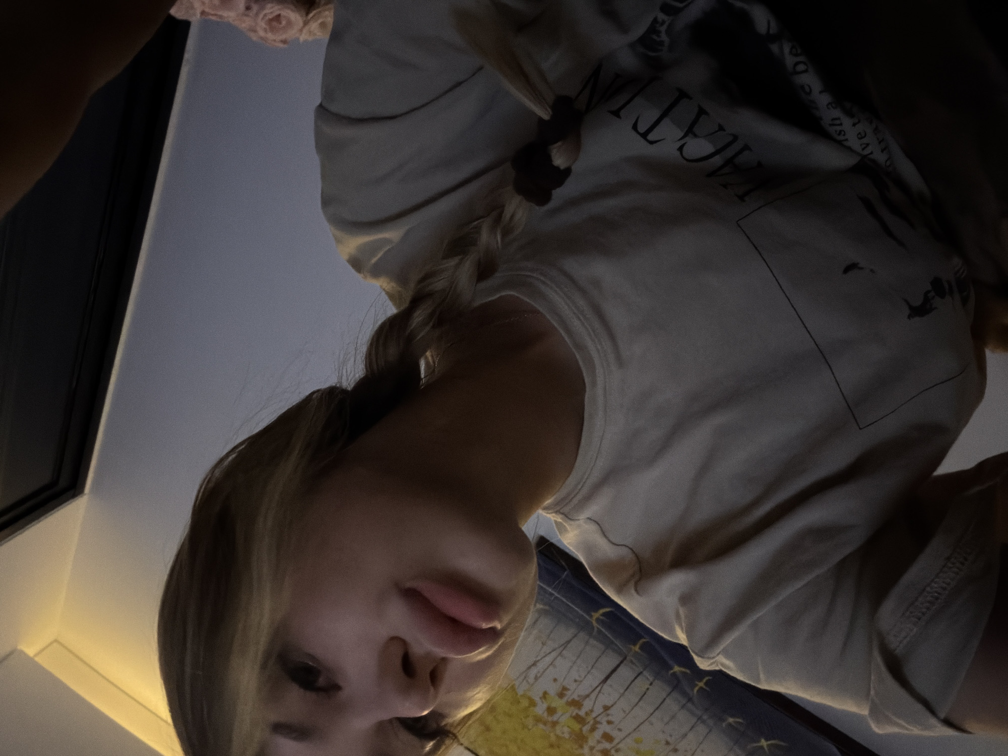 The image shows a young woman with blonde hair in a braid, wearing a light-colored t-shirt. She is looking towards the camera, with her lips slightly parted. Her hair is pulled back and braided, with a dark scrunchie holding it in place. The background is dimly lit, with a wall and what appears to be a framed artwork or poster above and behind her. The overall lighting suggests an indoor setting, possibly a bedroom or living area. There are no visible elements that place this image specifically in the city of Đà Nẵng, Vietnam.