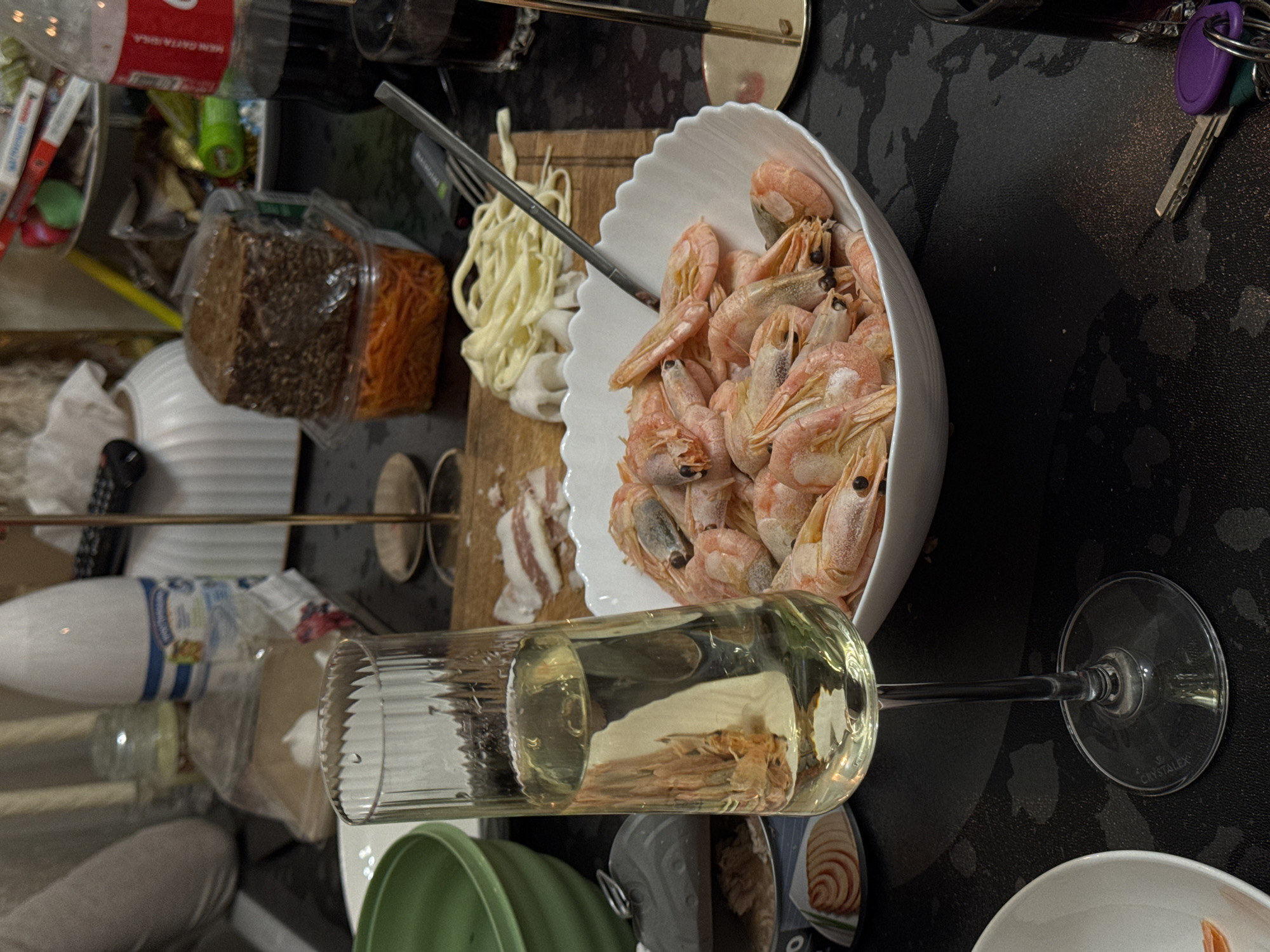 The image presents a cluttered table with various food items and objects, likely in Moscow, Russia. In a white scalloped bowl, a generous serving of cooked, unpeeled shrimp is displayed. Adjacent to the bowl, a wooden cutting board holds what appears to be fresh pasta, thin slices of meat, and a metal serving fork. Nearby are containers of dark bread and shredded carrots.
A glass of white wine is positioned prominently in the foreground. Other objects include a partially visible bottle of Coca-Cola, a jar, and a bottle of dairy product. Keys are present on the table, suggesting a casual, lived-in environment.
The table surface is black with a mottled texture. The overall impression is that of a meal preparation or the aftermath of a gathering. No people are fully visible, though a partial view of a person's clothing is at the bottom. The specific location within Moscow is not identifiable from the image.