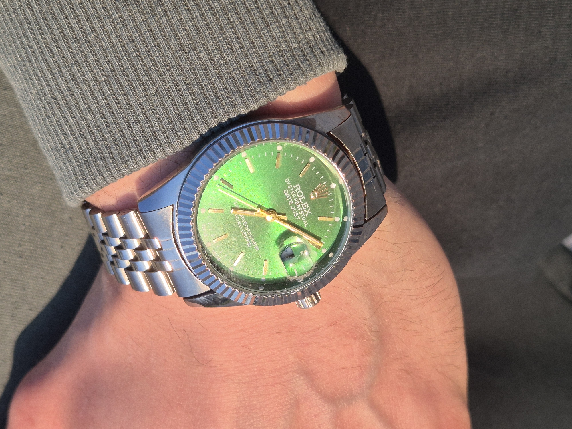 A wrist wearing a Rolex Datejust watch with a green dial is visible. The band is a silver jubilee style. The wrist is likely attached to an arm wearing a green knit sweater. The background is dark, suggesting indoor lighting conditions. The location is not discernable from the image.
