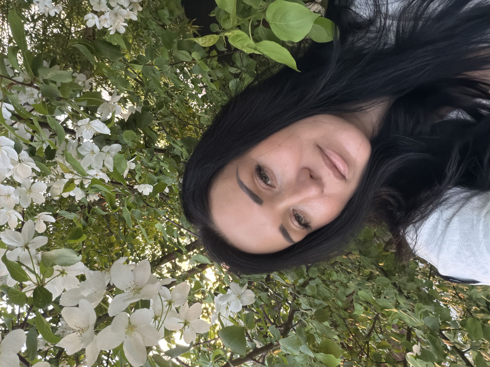 The image is a diagonally oriented outdoor portrait featuring a woman with long, dark hair, dark eyes, and well-defined eyebrows. Her face is angled towards the viewer, displaying a serene and gentle expression with slightly parted lips. A light-colored garment is partially visible on the right side of the frame.
The woman is positioned amidst a dense natural backdrop of green leaves and numerous white blossoms, likely from a flowering tree such as an apple or cherry. These flowers are in full bloom, indicating a setting during spring or early summer. Bright, natural daylight filters through the foliage, creating a luminous effect and suggesting a pleasant, clear day. The perspective looks upwards into the tree canopy, with the woman's face framed by the surrounding blossoms and leaves. No text is visible. Based on the provided context, this scene is set in Dzerzhinsk, Russia.